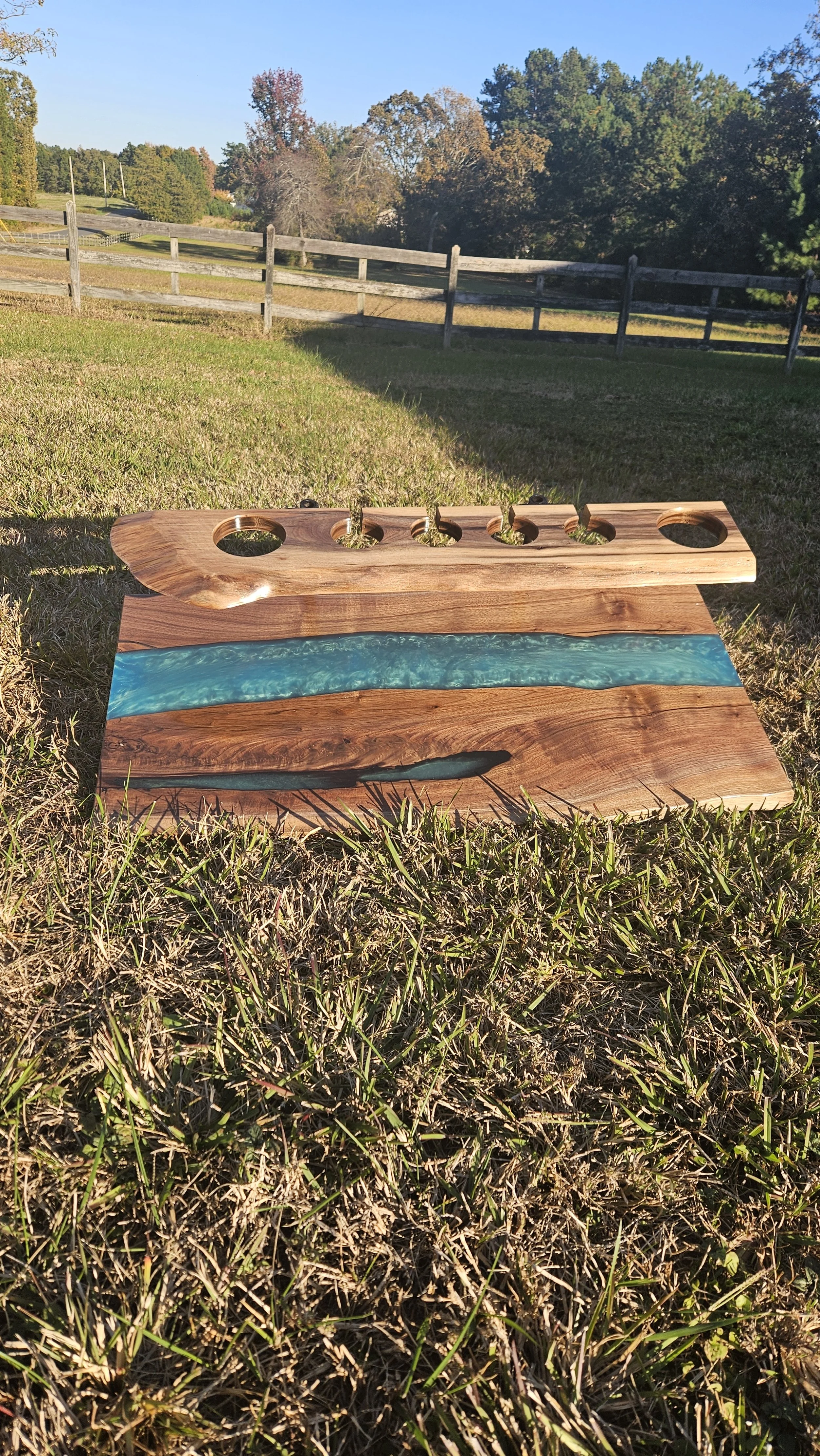 A wooden art piece with a blue resin river design resting on grass with trees and a wooden fence in the background.