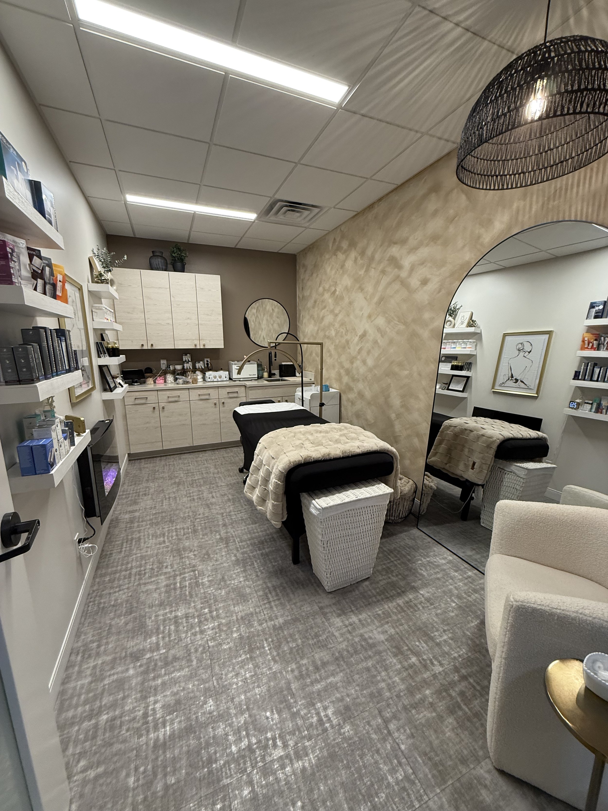 A spa or massage therapy room with a massage table, a large mirror, and shelves filled with skincare products, decorated in neutral tones.
