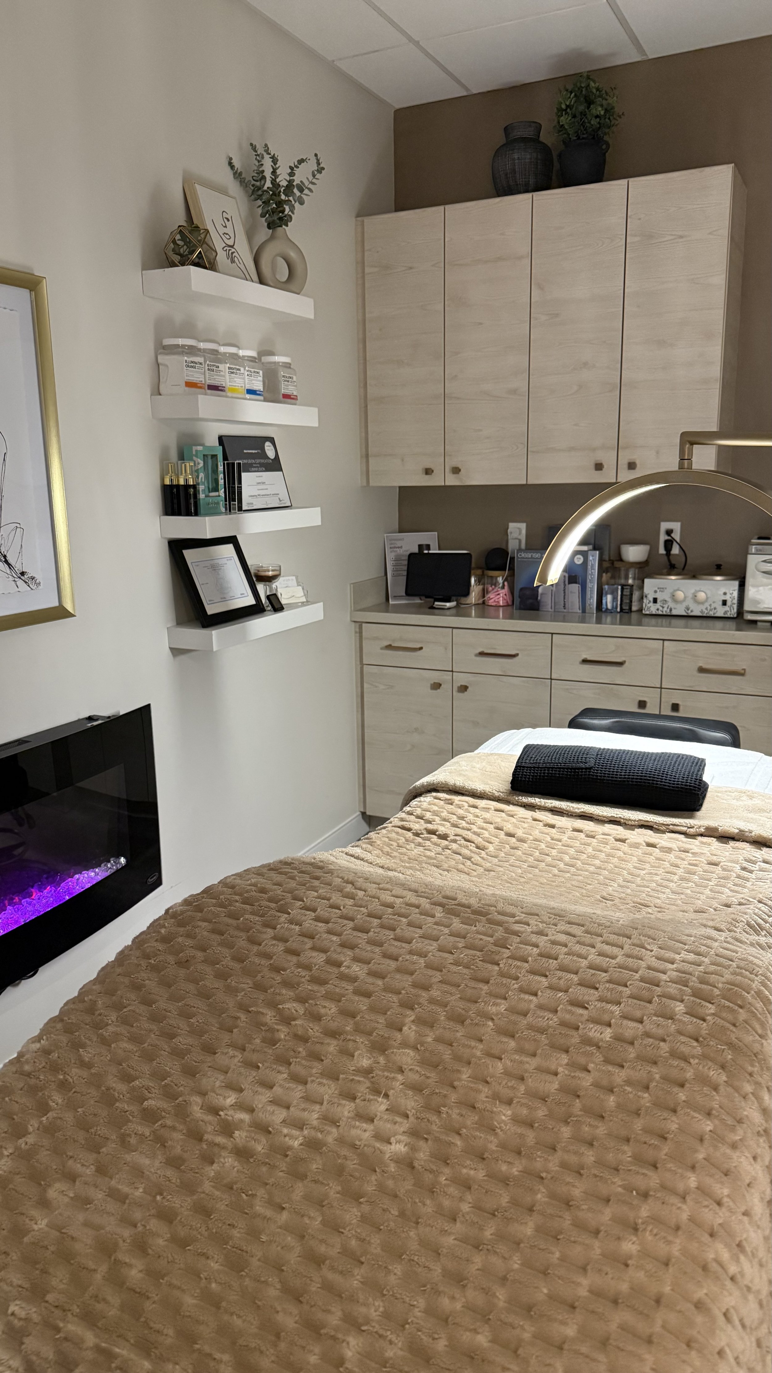 A massage therapy room with a massage table covered in a tan textured blanket, a black towel on top, wall-mounted shelves with skincare products, a framed certificate, and a small electronic device on a beige cabinet. There is a black electric firepl