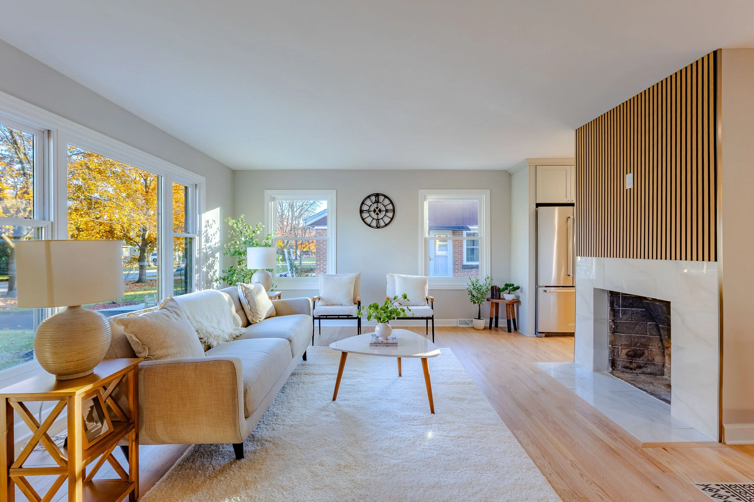 Fox Point, WI - Real Estate - Bright, modern living room with large windows, white sofa, wooden side table, coffee table, armchairs, fireplace with wooden panel above, hardwood floors, and some indoor plants.