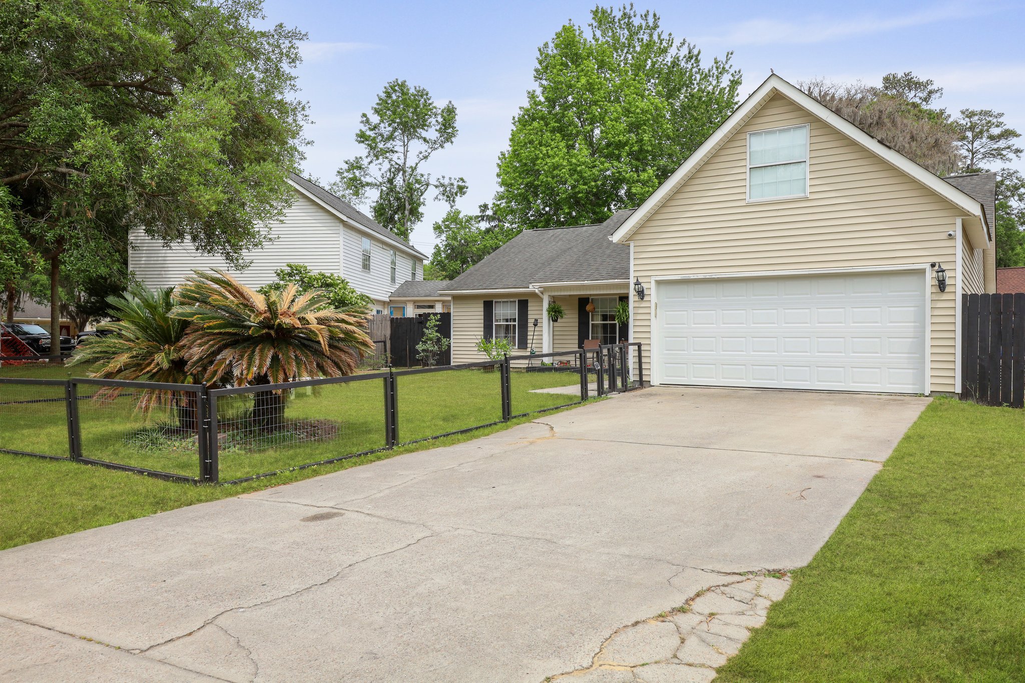 Residential house with beige siding, white garage door, and front yard with a small tree and plants, surrounded by a black fence, on a driveway.