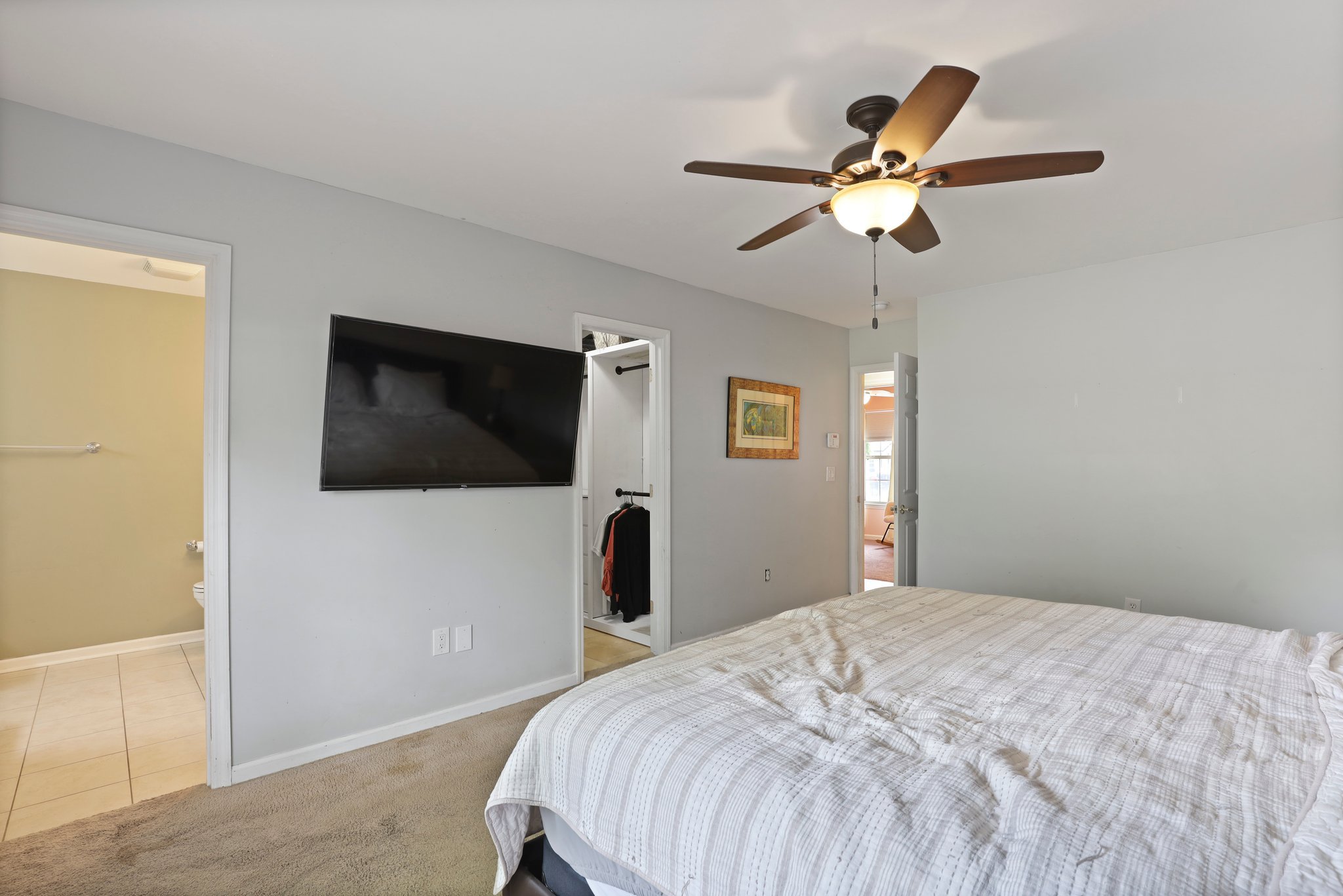 Bedroom with ceiling fan, flat-screen TV, bed with striped bedding, two open doors leading to a bathroom and closet, light gray walls, and a framed picture on the wall.