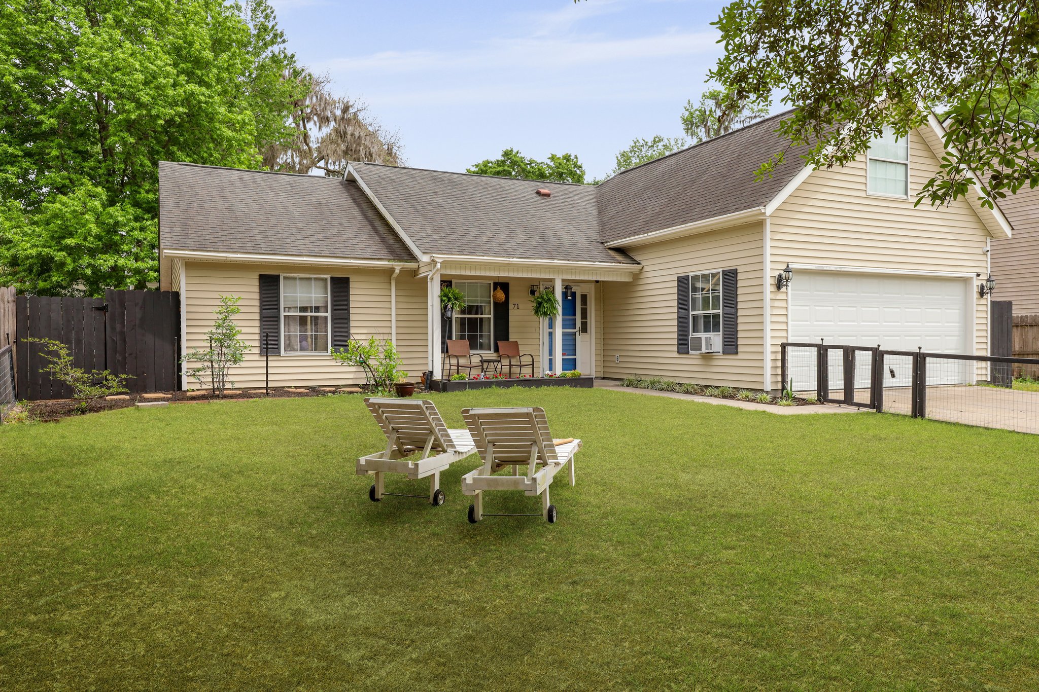 Single-story beige house with a front porch, green lawn, two lounge chairs, and a double garage. Surrounded by a wooden fence and trees.