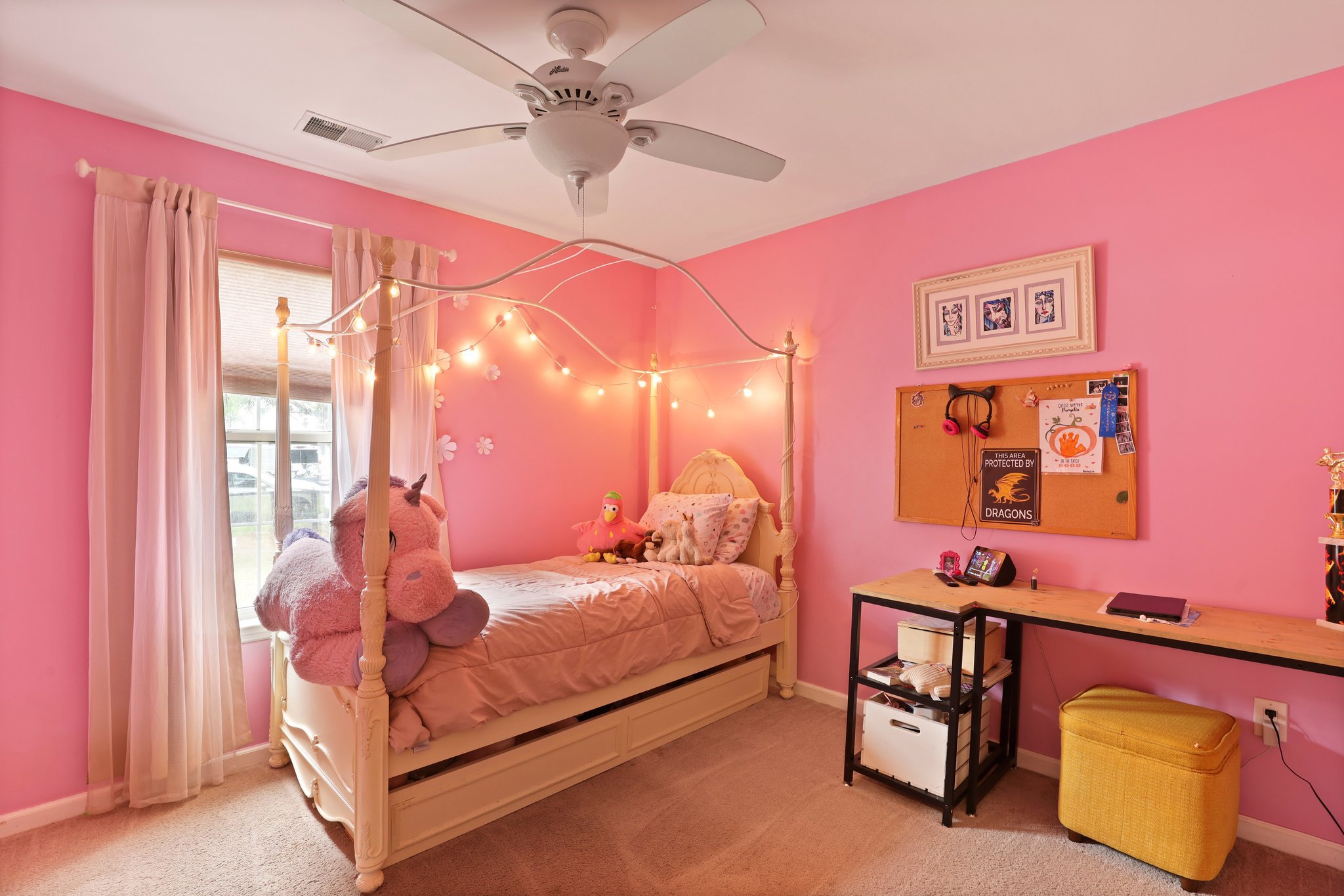Pink-themed child’s bedroom with a canopy bed, string lights, stuffed toys, and a desk. Walls are decorated with framed pictures and a corkboard. The room features a ceiling fan and a window with sheer curtains.