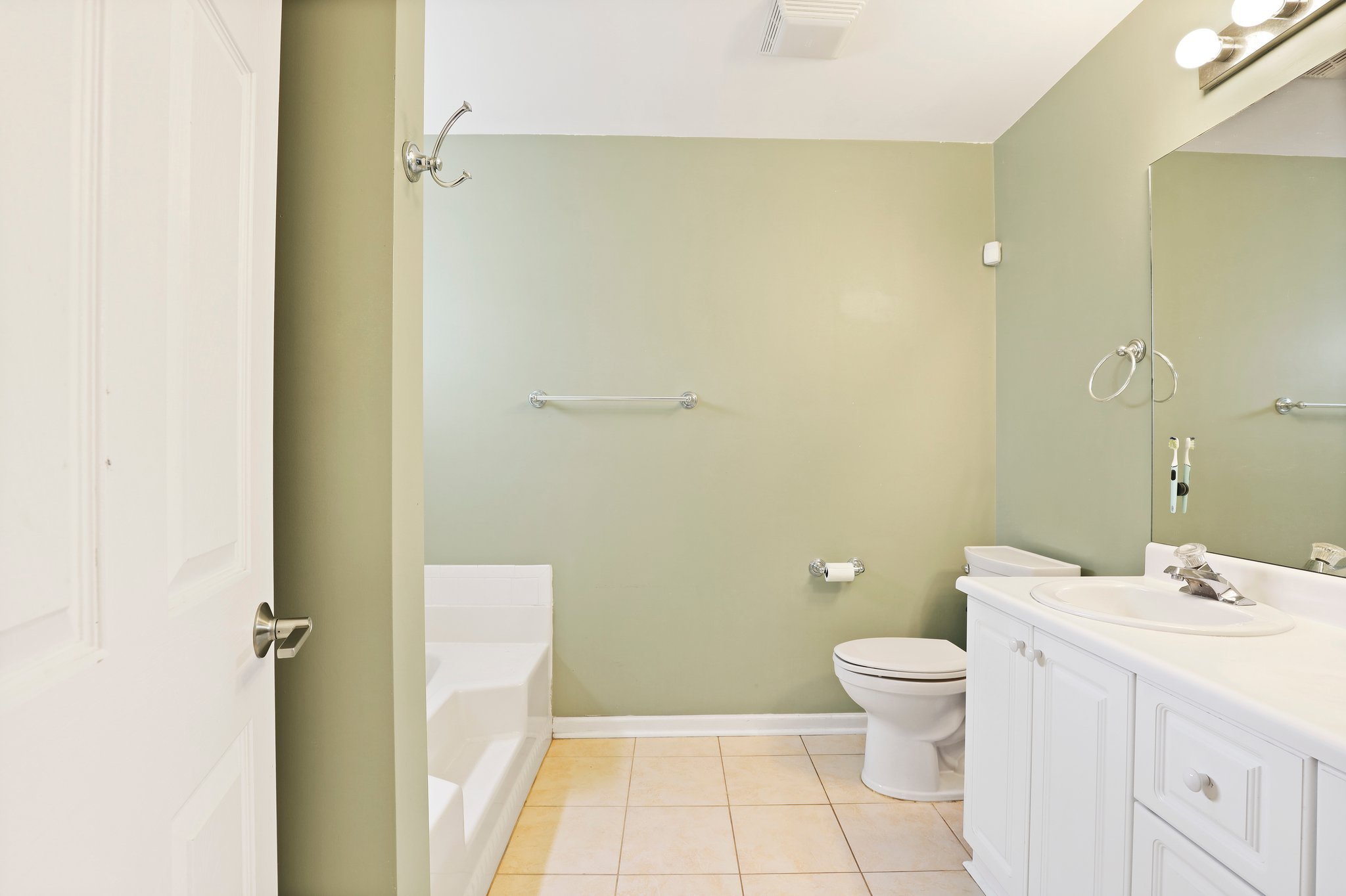 Bathroom with green walls, tan floor tiles, a white vanity with sink, a mirror, a toilet, and a tub. The room has a towel rack, a toilet paper holder, and ceiling lights.