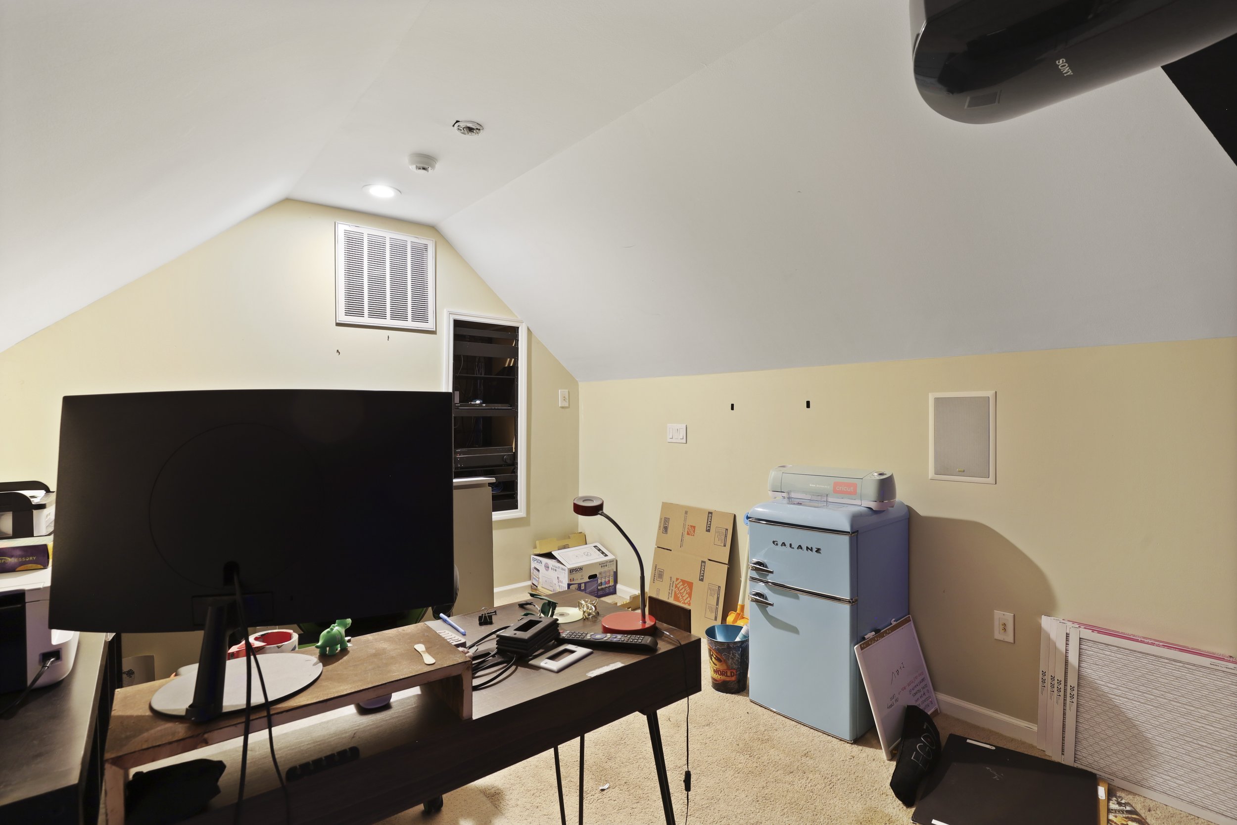 Home office in attic with a sloped ceiling, featuring a desk with a computer monitor, a small retro-style light blue refrigerator, various electronics, boxes, and storage items.