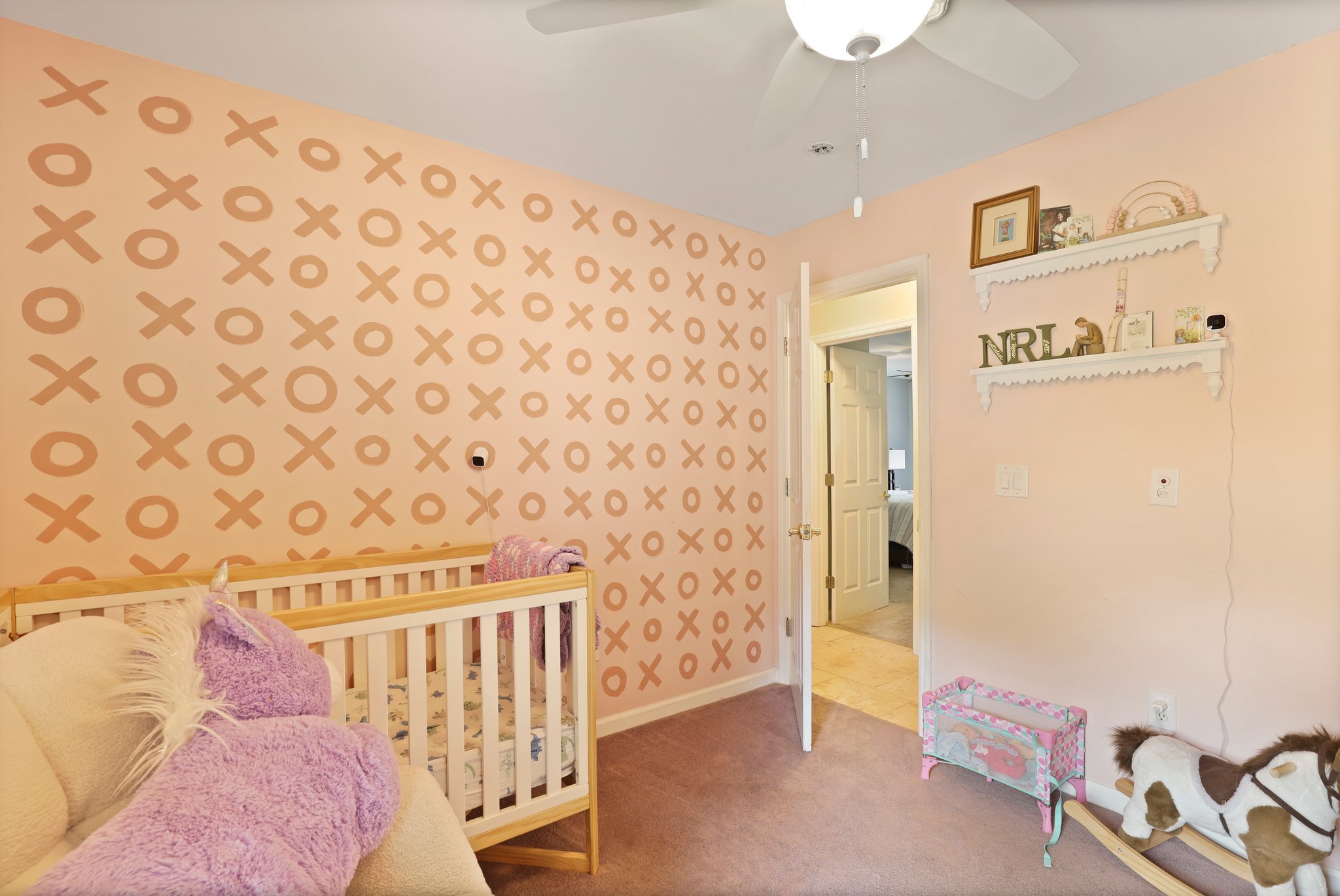 A nursery room with a pink XO patterned wall, a wooden crib, a plush unicorn, and white shelves with decorative items. A ceiling fan with a light is on the ceiling, and an open door leads to another room.
