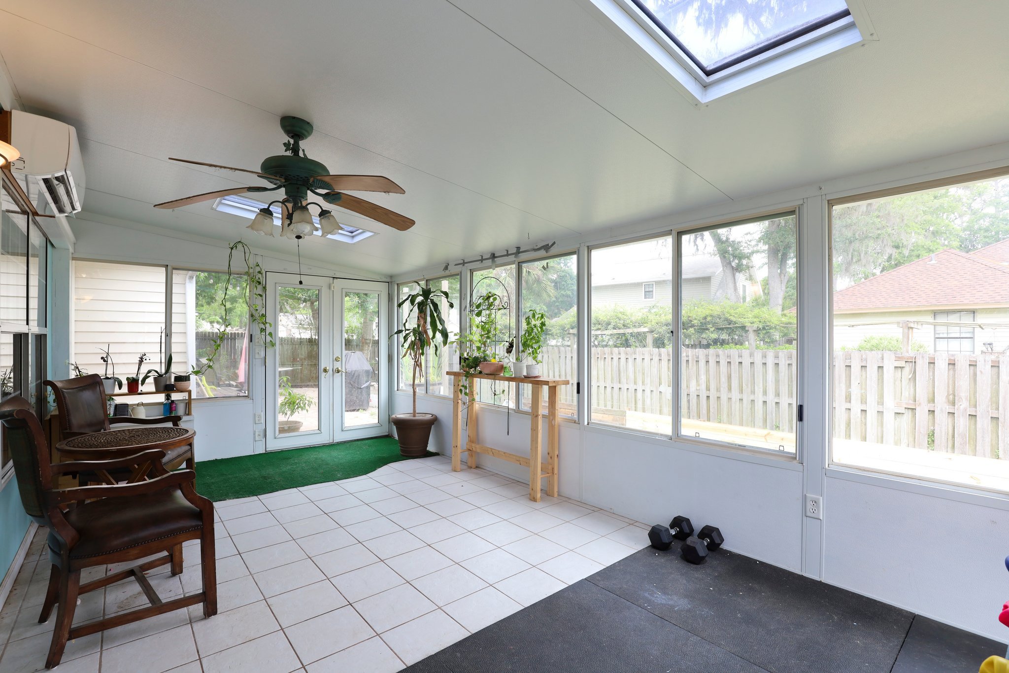 A sunroom with large windows, a skylight, tile flooring, and a ceiling fan. There are potted plants, a small table with chairs, and dumbbells on the floor. A view of a backyard with a wooden fence is visible through the windows.