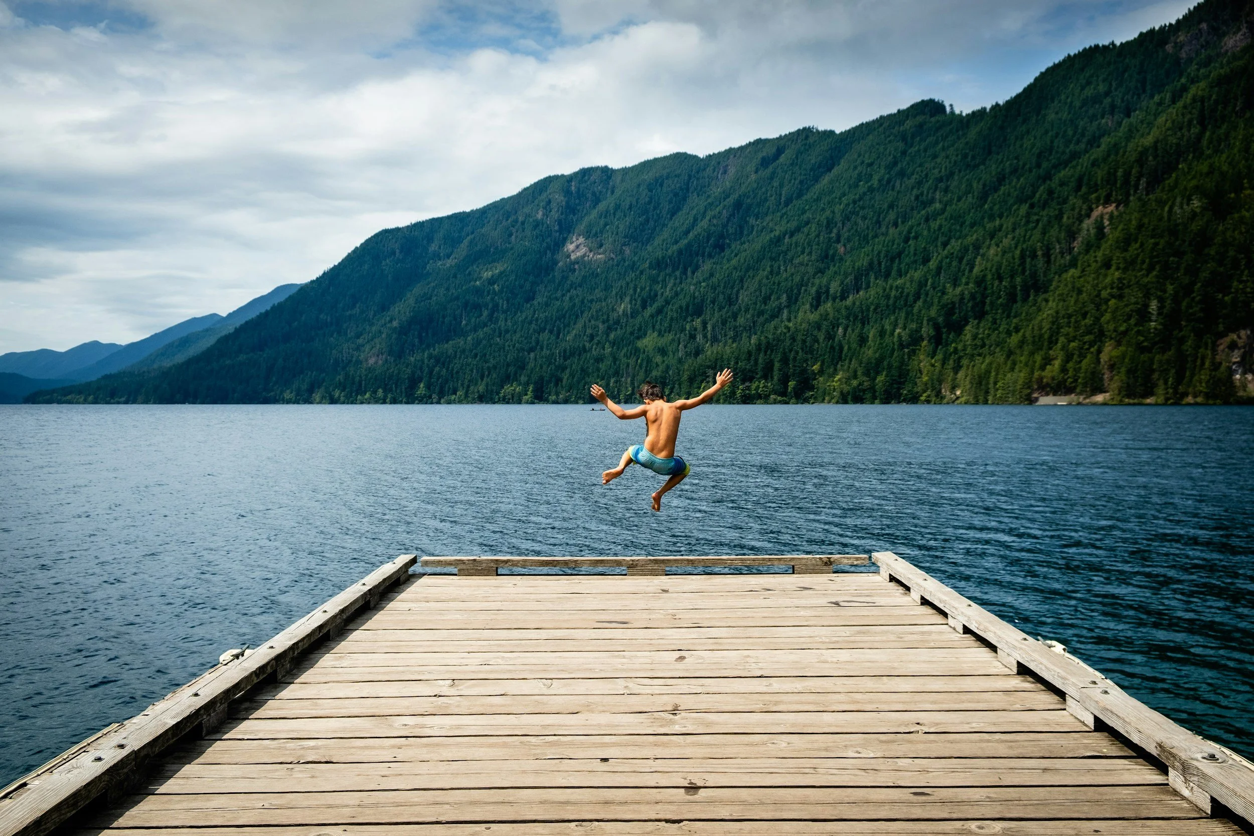 A boy in blue swim trunks jumping off a wooden dock into a lake, with mountains and trees in the background under a partly cloudy sky.