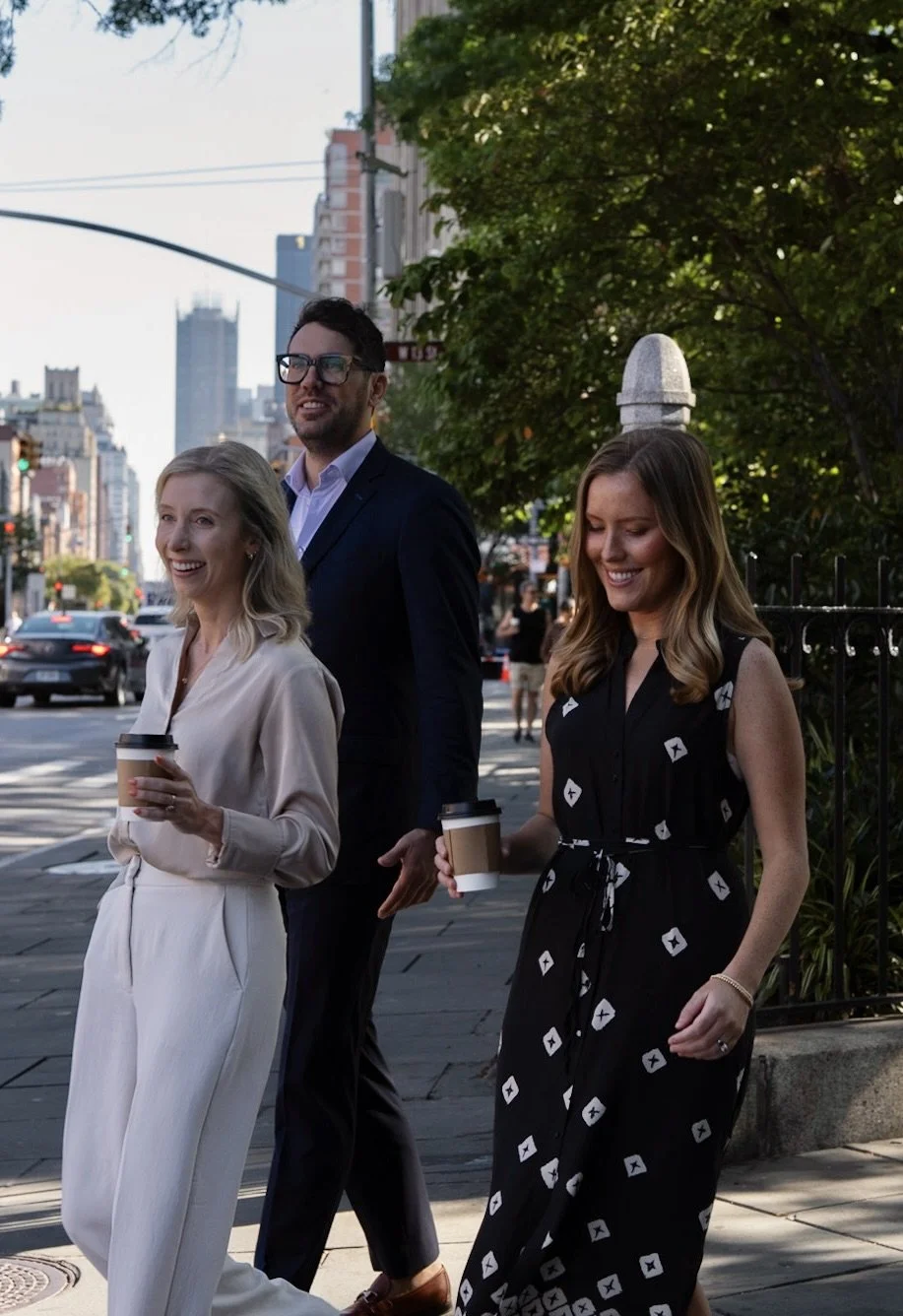 Peltoma Capital Partners advisor team walking on a New York city sidewalk, smiling and holding to-go coffee cups, with buildings and trees in the background.
