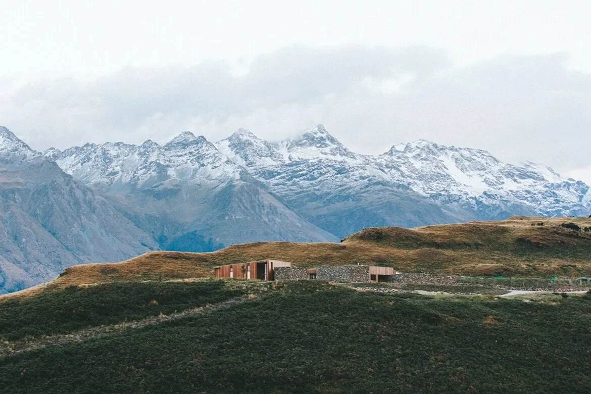 A mountain landscape with snow-capped peaks in the background and a grassy hill with modern buildings in the foreground.