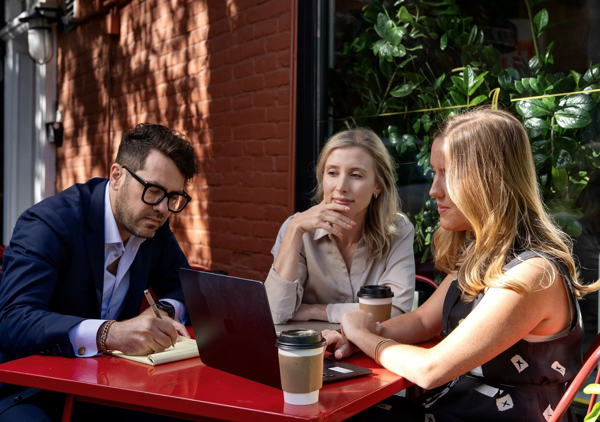 Peltoma Capital Partners advisor team sitting around a red table outdoors, engaged in discussion. Coffee cups and a laptop are on the table. Behind them, there is a brick wall and green foliage.
