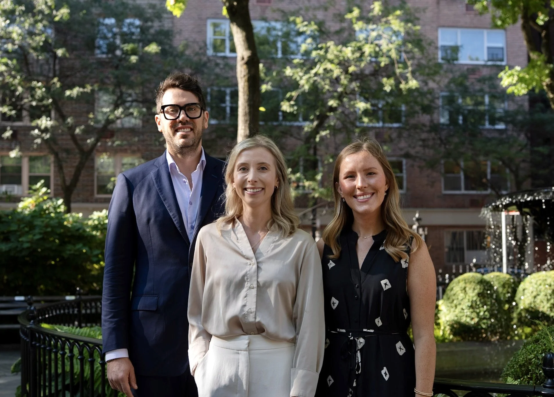 Peltoma Capital Partners advisor team standing outdoors in a New York City park with greenery and trees, in front of a brick residential building.