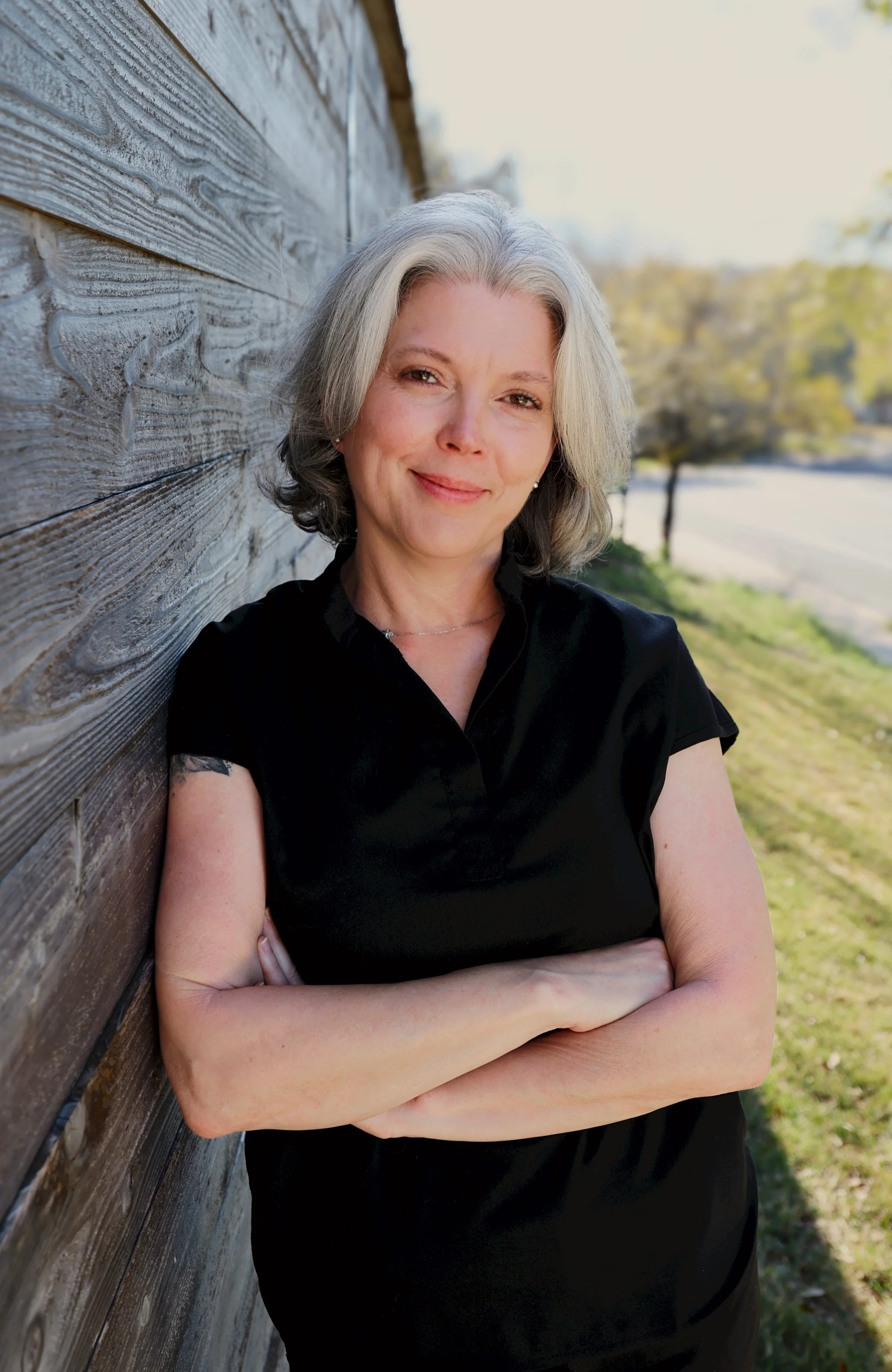 A middle-aged woman with gray hair leaning against a wooden wall outdoors with arms crossed, smiling at the camera.