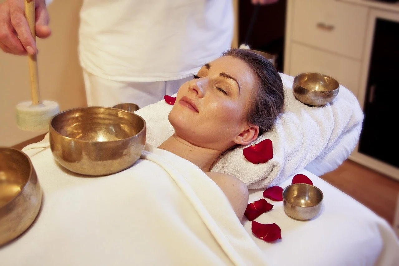Woman receiving a sound therapy or healing treatment with singing bowls on a massage table, surrounded by rose petals.