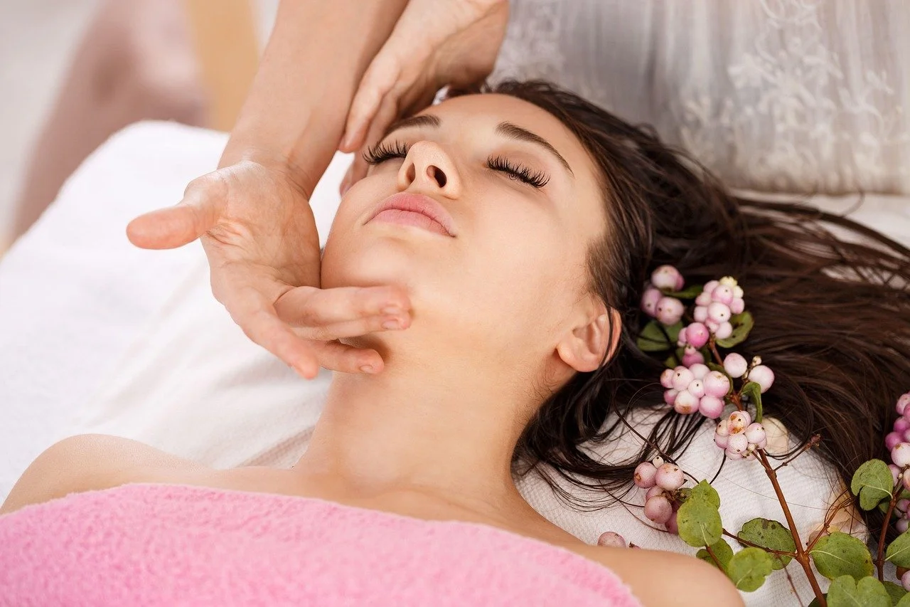 A woman receiving a facial massage while lying down with pink flowers in her hair.
