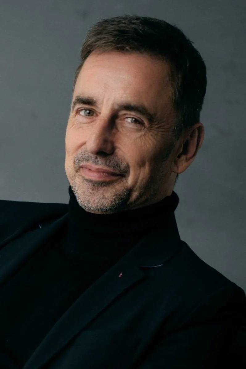 Tom is smiling into the camera and wearing a dark roll neck jumper