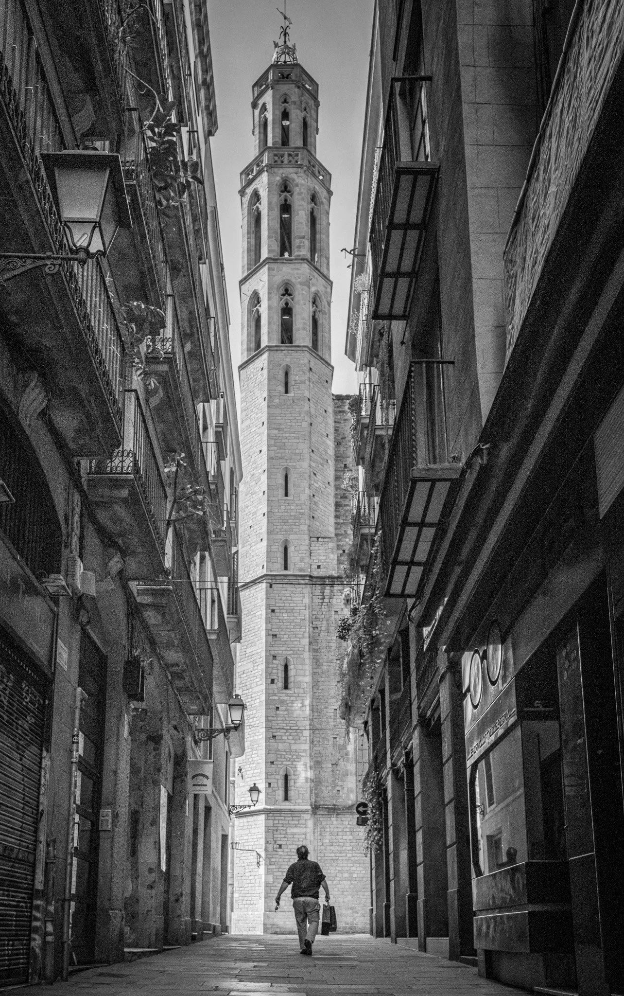 Black and white street photograph of a narrow Barcelona alley leading toward a tall church bell tower, with a person walking in the distance.