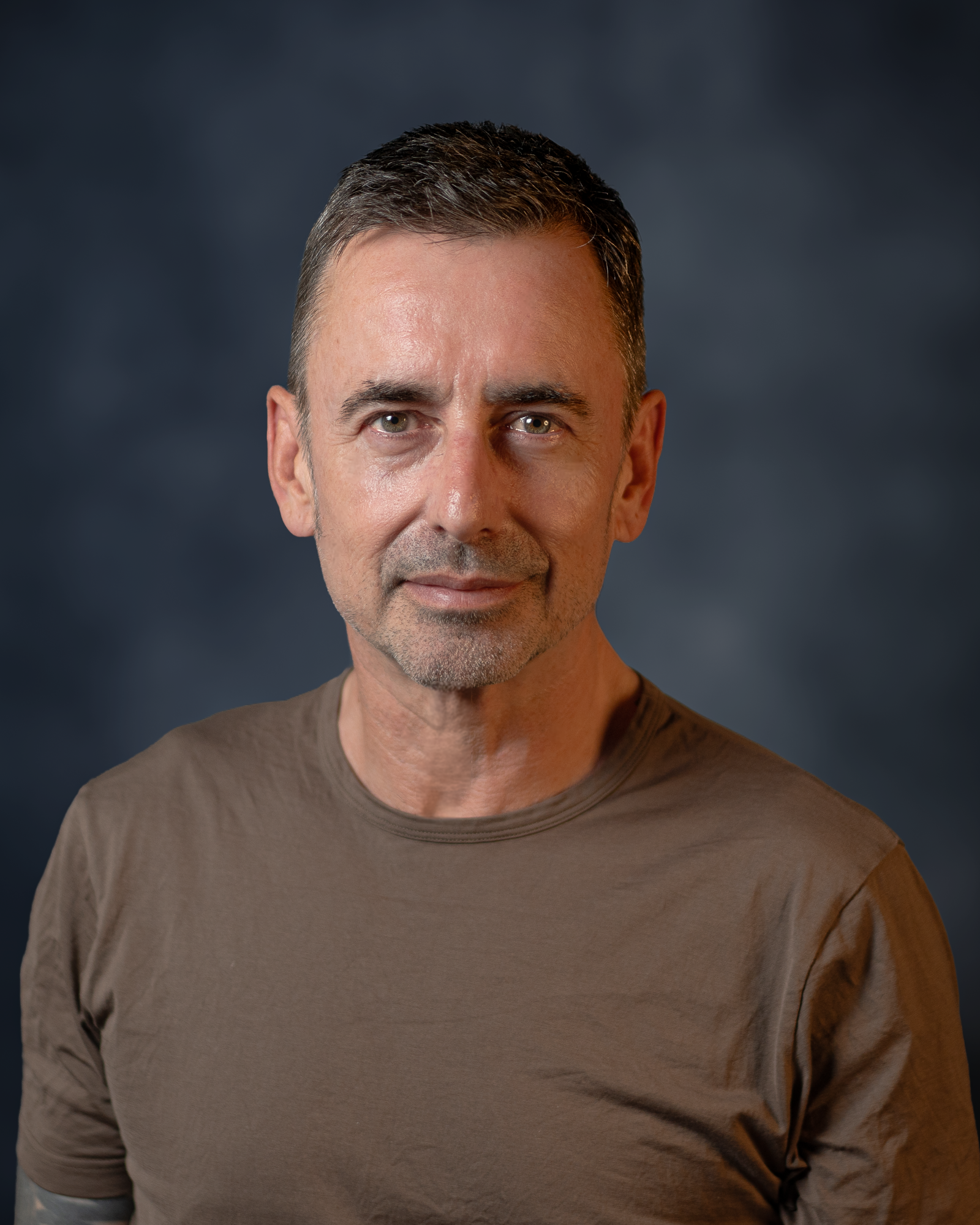 Portrait of Tom Beardshaw, executive coach, in a brown shirt against a dark blue-grey background, looking directly at the camera with a calm expression.