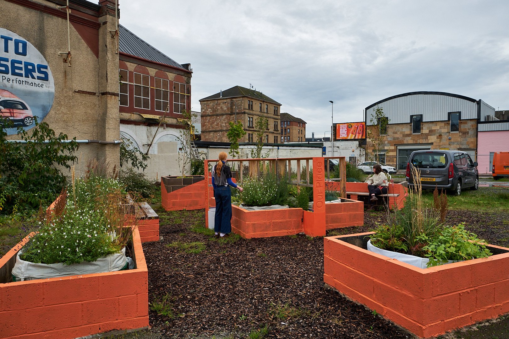 A community garden with orange planter boxes and people working and sitting. Background includes buildings, a sign, and parked vehicles. Overcast sky.