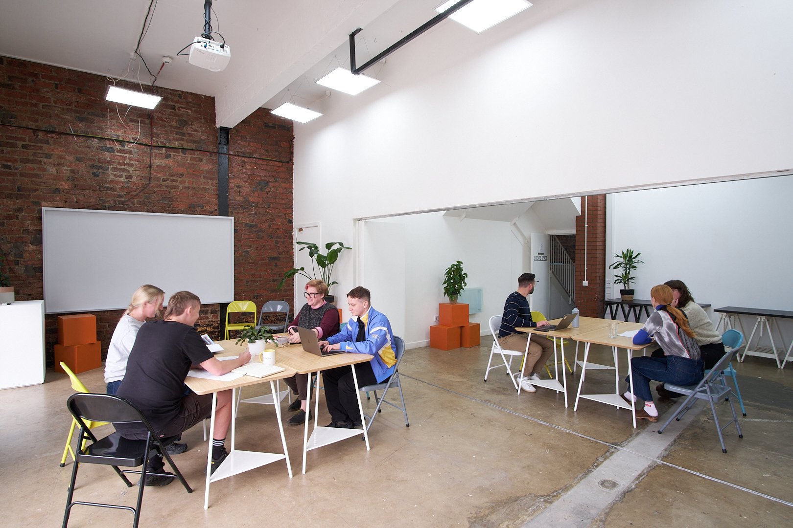 Modern coworking space with people working at tables, exposed brick wall, and indoor plants.