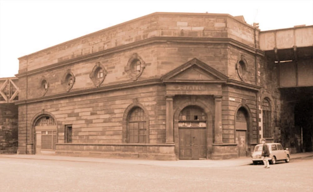 Sepia-toned image of an old stone building with arched windows and doors, adjacent to a bridge structure. A vintage car is parked nearby.