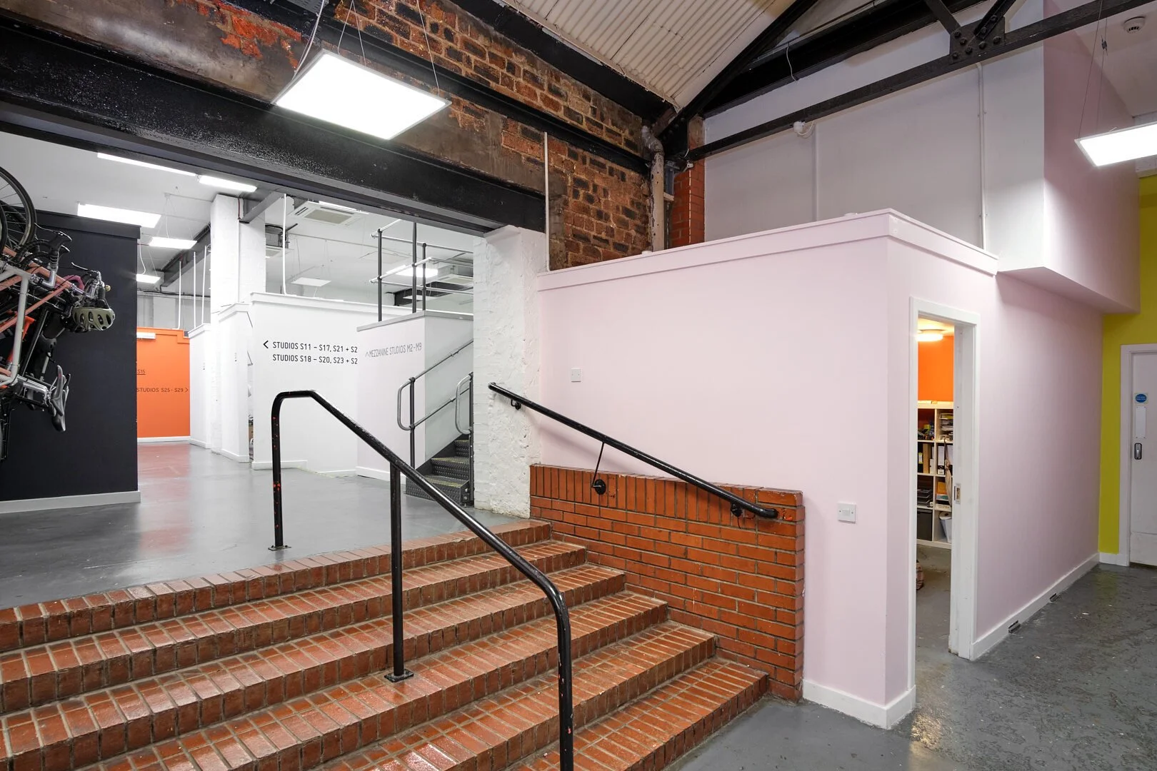 Interior of a modern workspace with brick stairs, pink walls, and signage directing to different studio spaces.