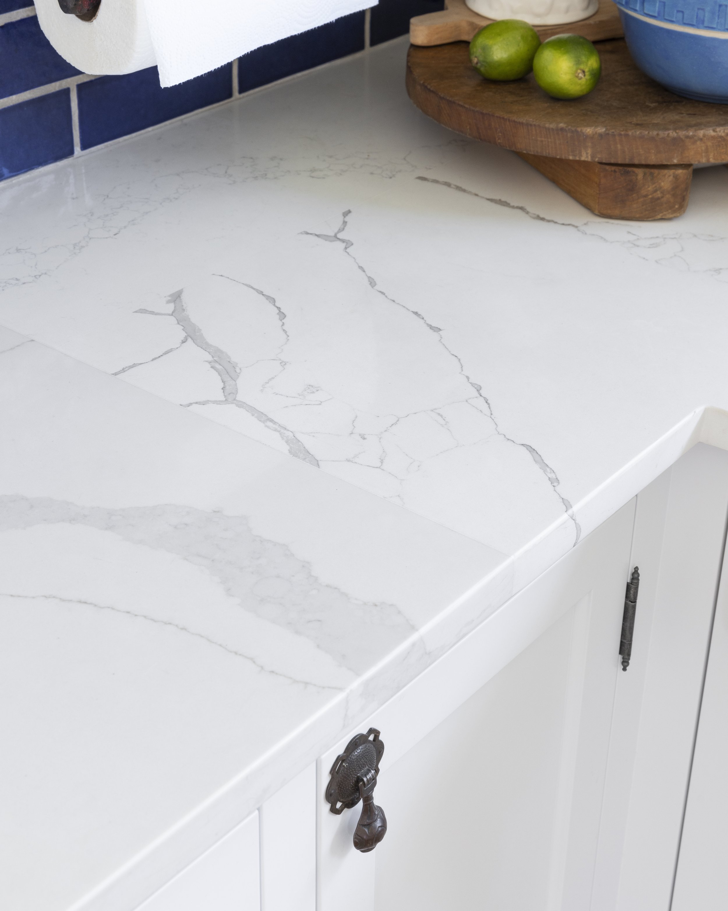 White marble kitchen countertop with blue backsplash, paper towel roll, wooden tray, blue bowls, and limes.