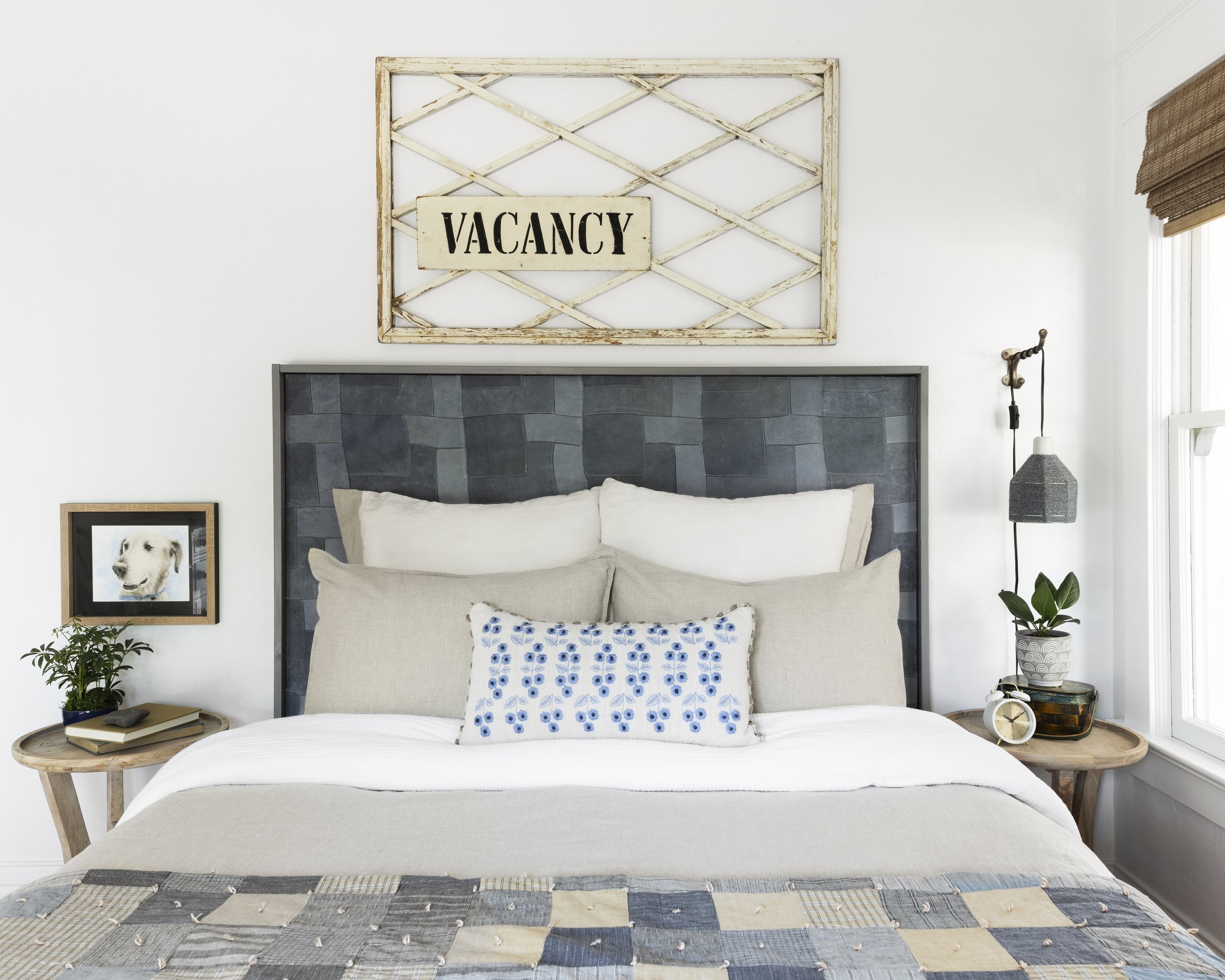 Bedroom with white and gray bedding, decorative 'Vacancy' sign above headboard, wooden side tables, framed dog photo, potted plants, and a wall-mounted lamp.