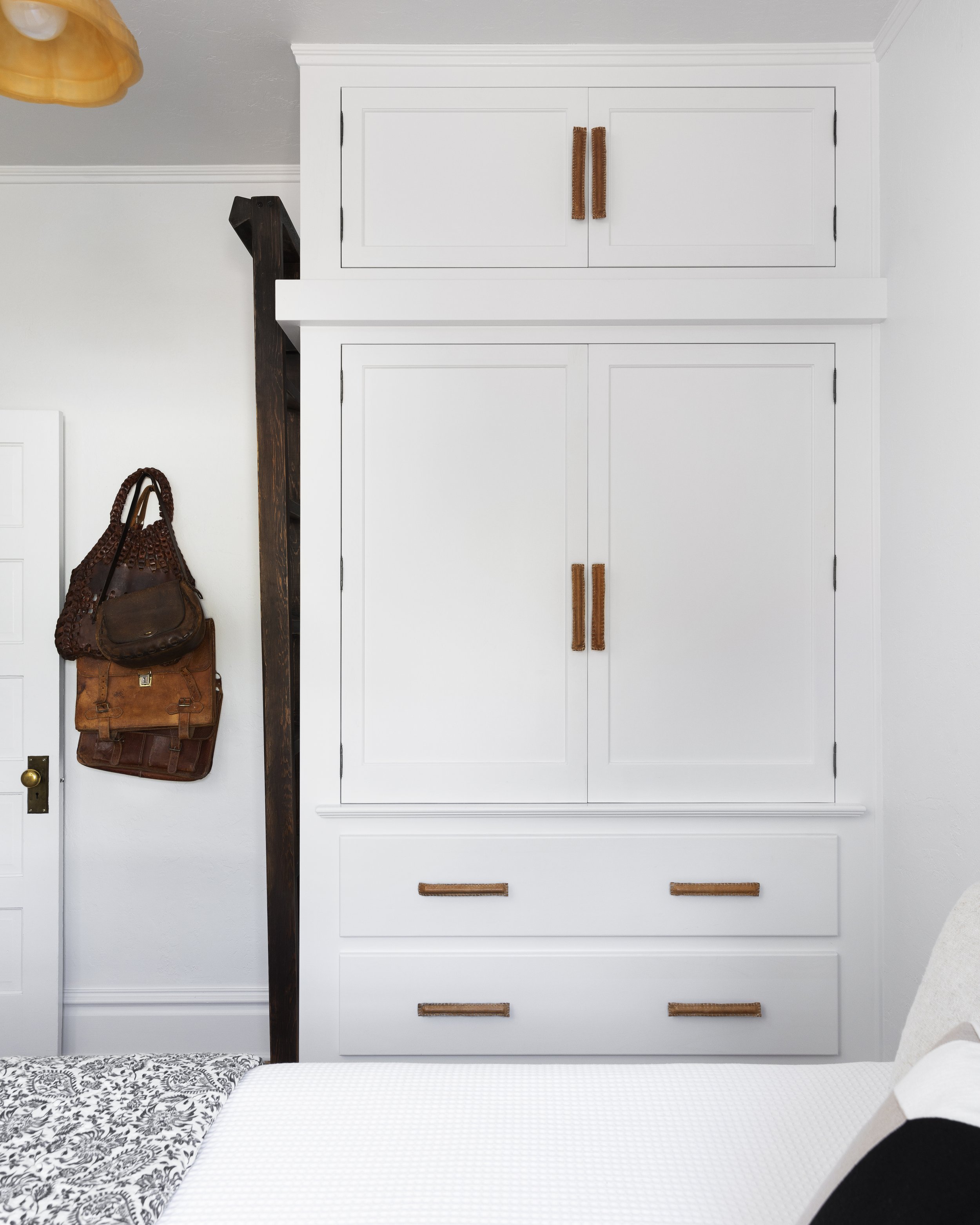 White built-in cabinet with leather handles, wooden beam, hanging brown leather bags, and patterned bedspread in a white bedroom.