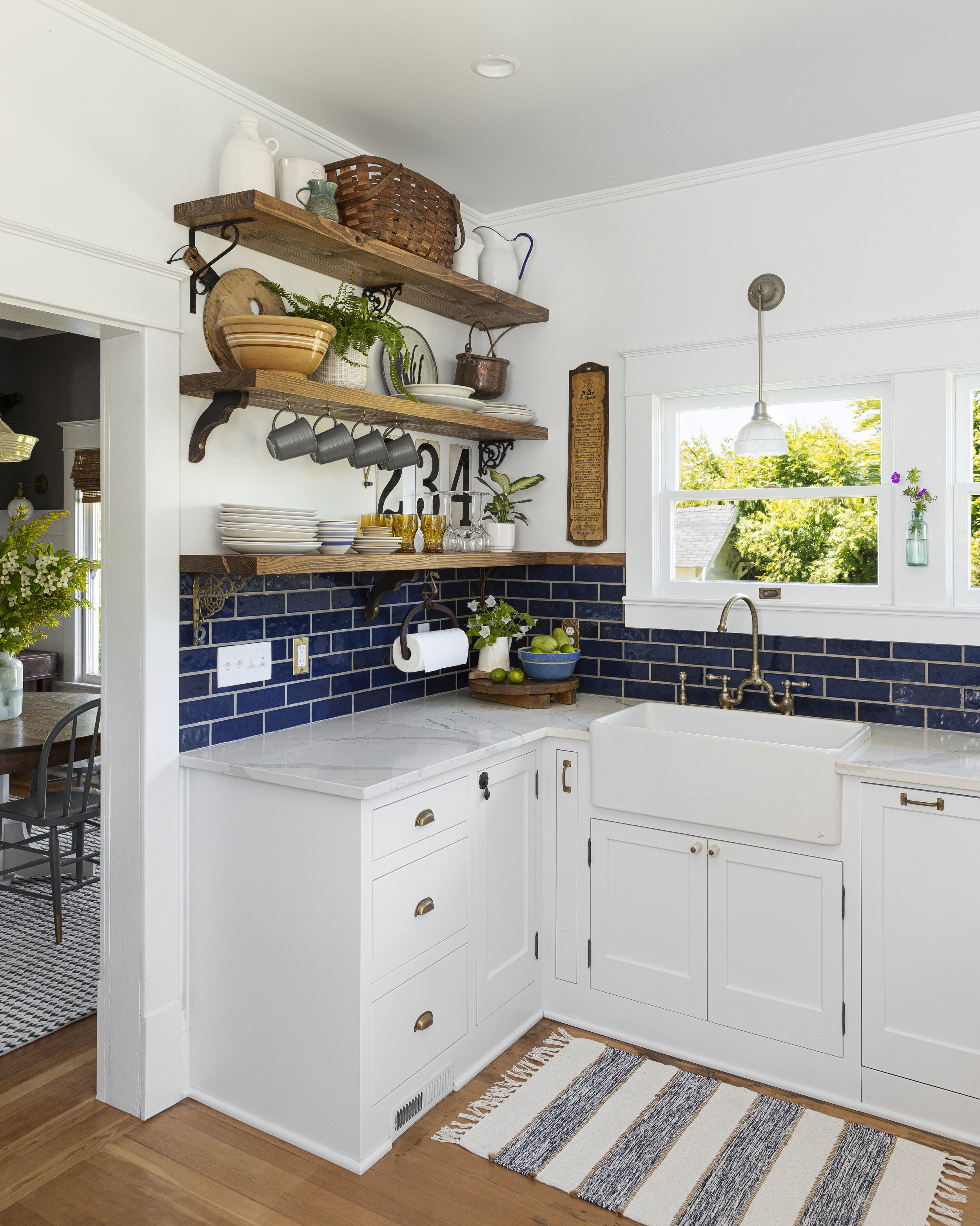 White kitchen with farmhouse sink, white cabinets, and blue subway tile backsplash. Wooden shelves hold dishes, baskets, and a plant. A pendant light hangs over the sink, and a striped rug is on the floor.