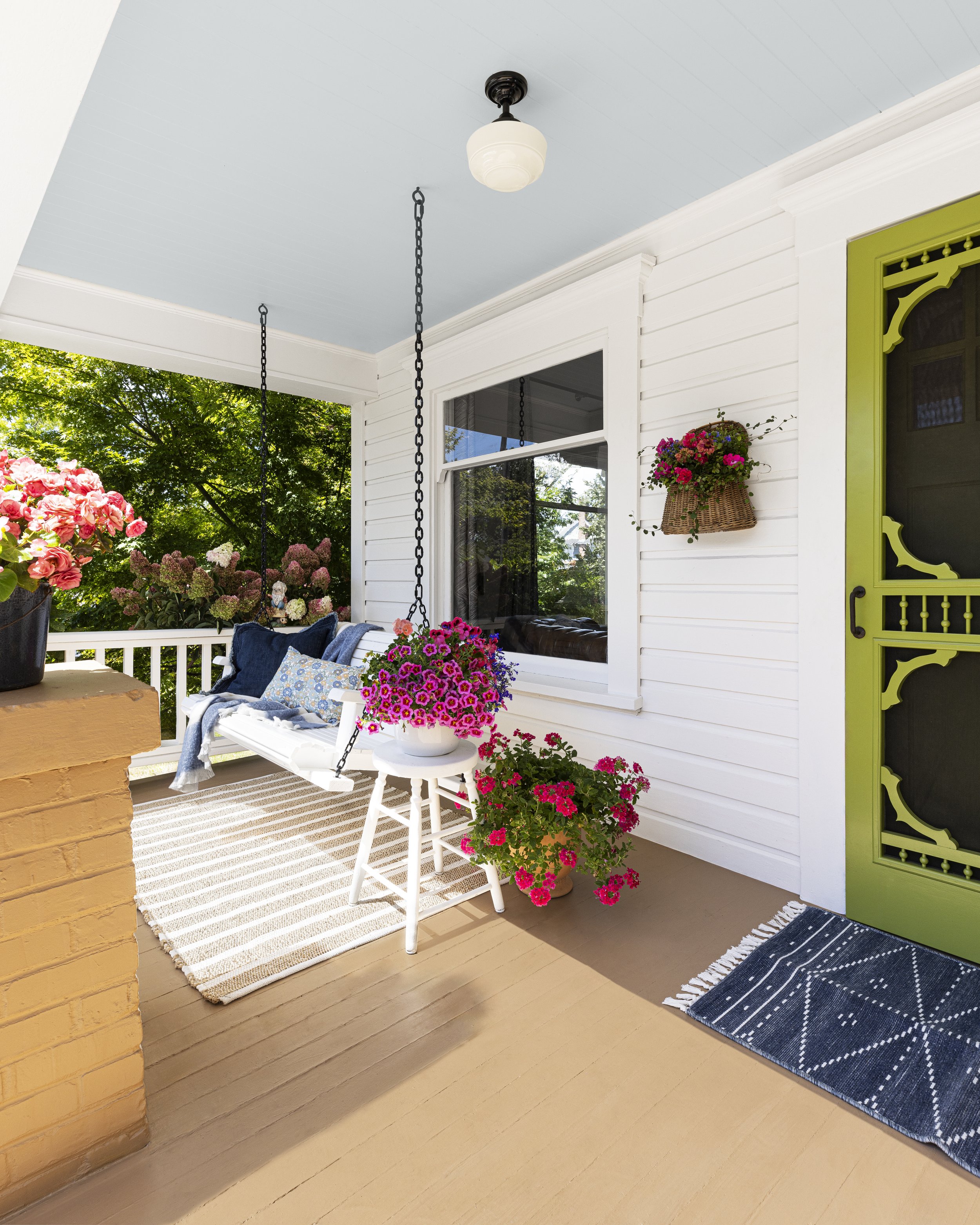 Front porch with a swing, colorful flowers, striped rug, wicker basket with flowers, and a green door with decorative design.