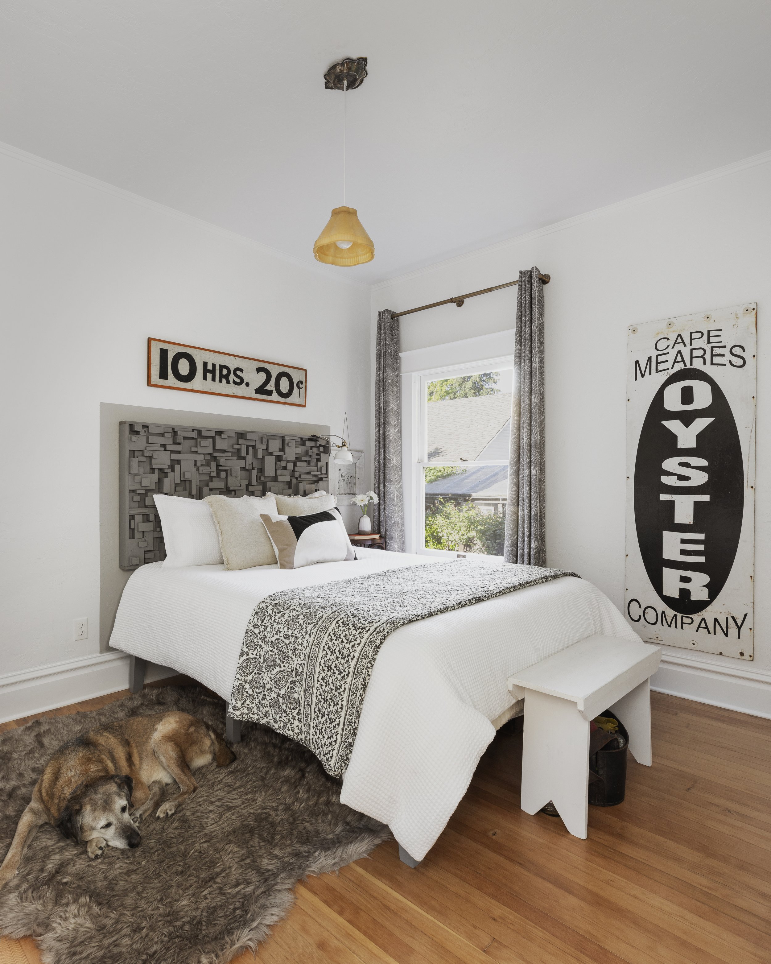 A cozy bedroom with a bed featuring white and patterned bedding, decorative pillows, and a textured headboard. There is a rug with a dog lying on it, wooden floors, and vintage signs on the walls. A window with curtains lets in natural light, and the