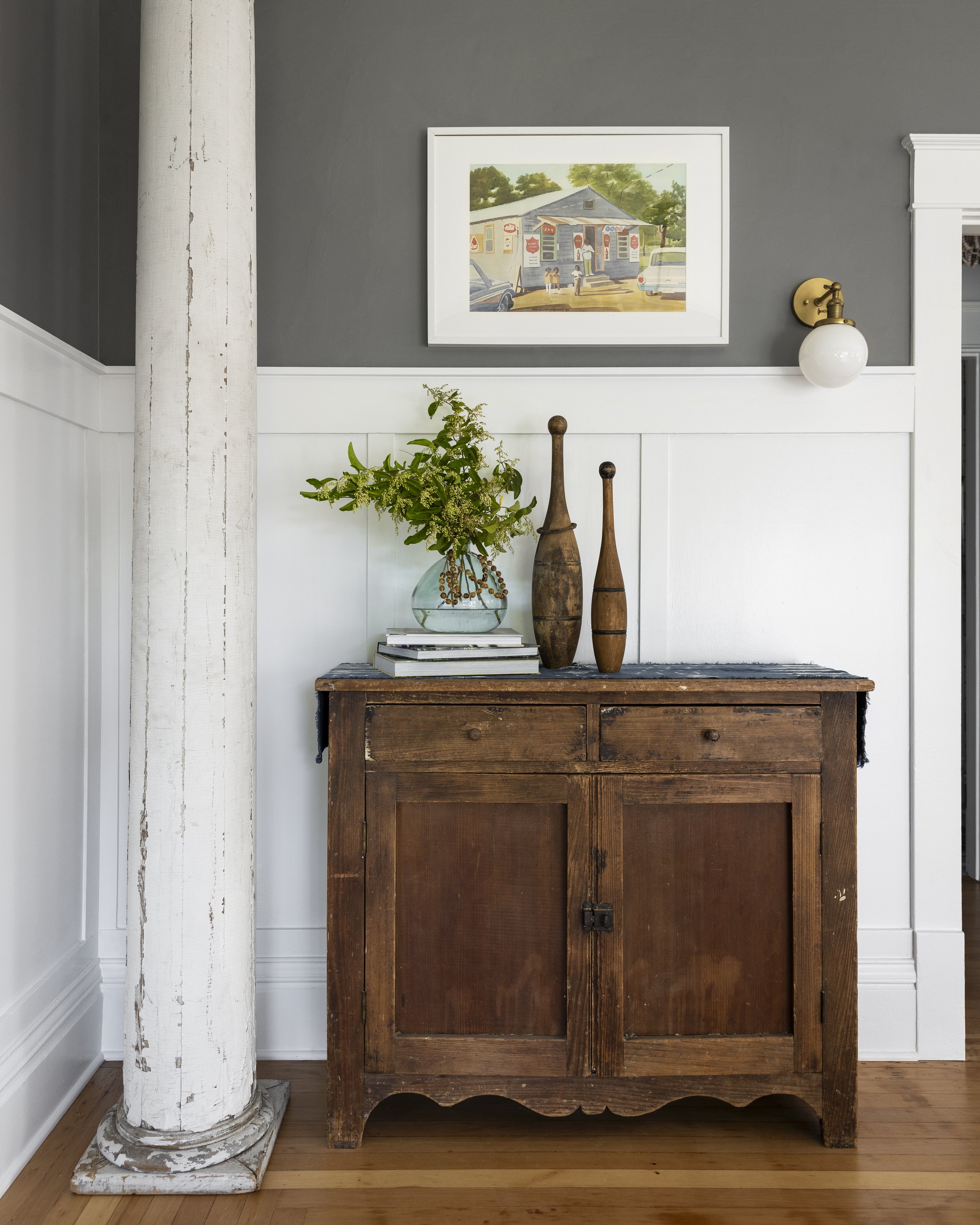 Room interior with a rustic wooden cabinet, decorative vase with greenery, two wooden sculptures, a framed painting, a white column, gray wall, white wainscoting, and hardwood floor.