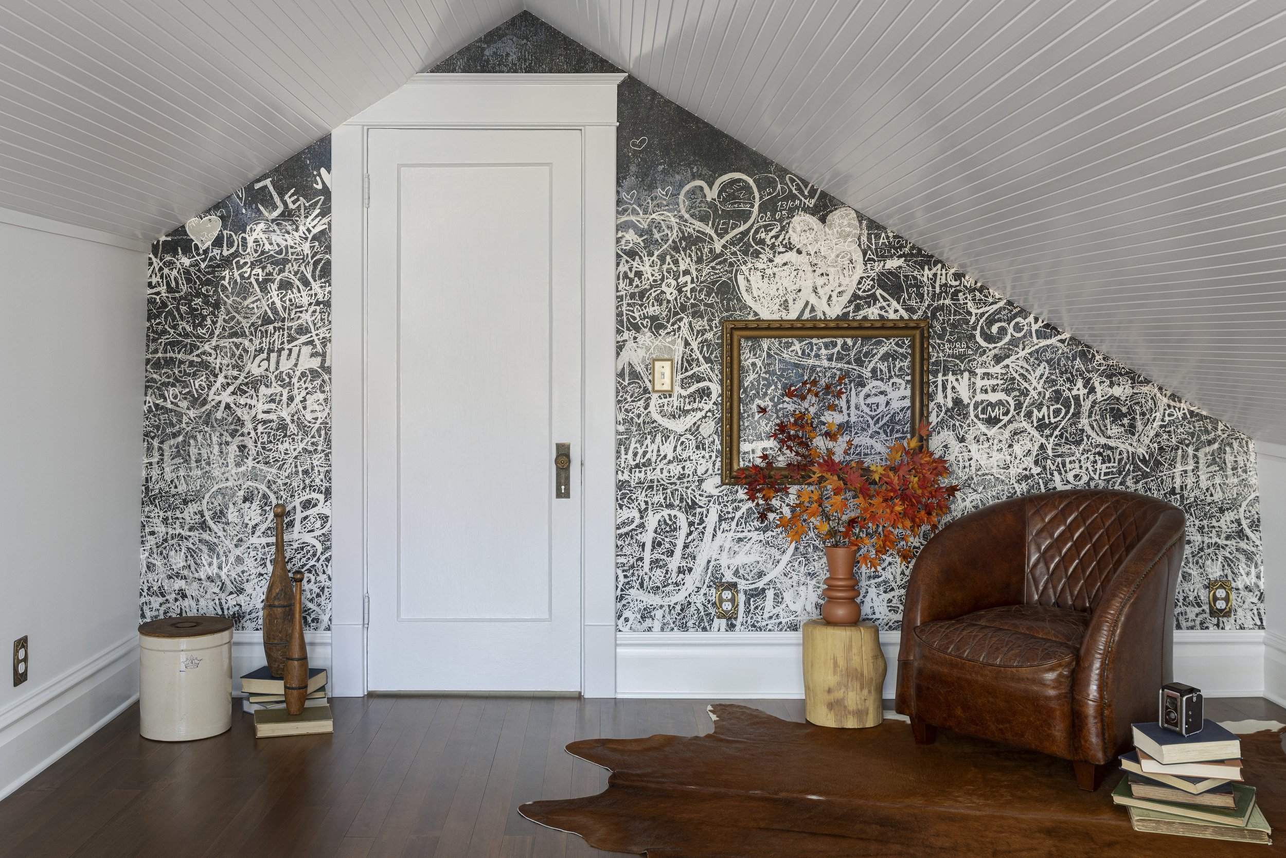 Attic room with white door, black graffitied wall, brown leather chair, small table with autumn leaves in a vase, books, and wooden decorative items on a wooden floor.