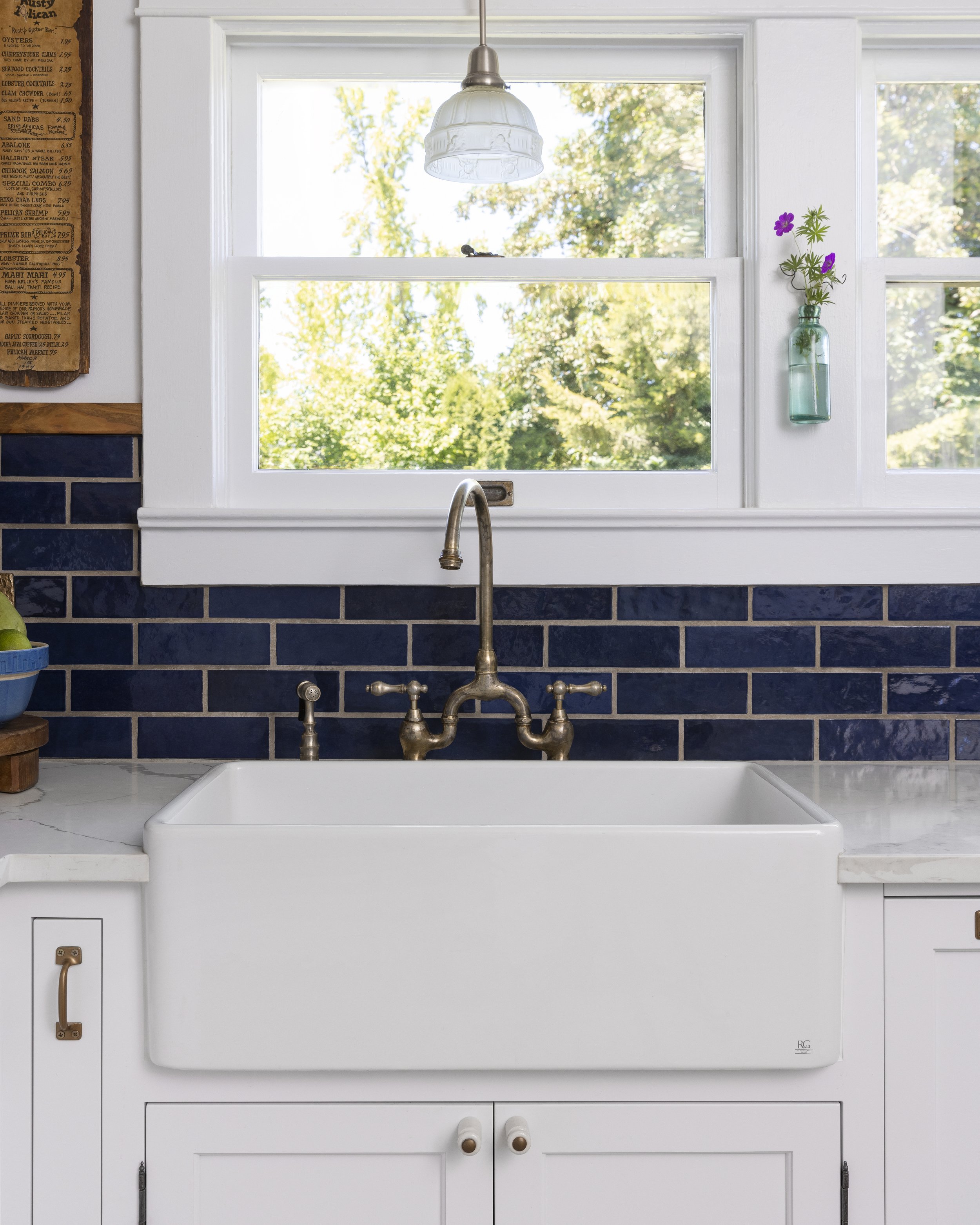 Farmhouse kitchen sink with brass faucet, blue tile backsplash, and a window with a view of trees. A hanging light fixture and a small vase with flowers are decoration.