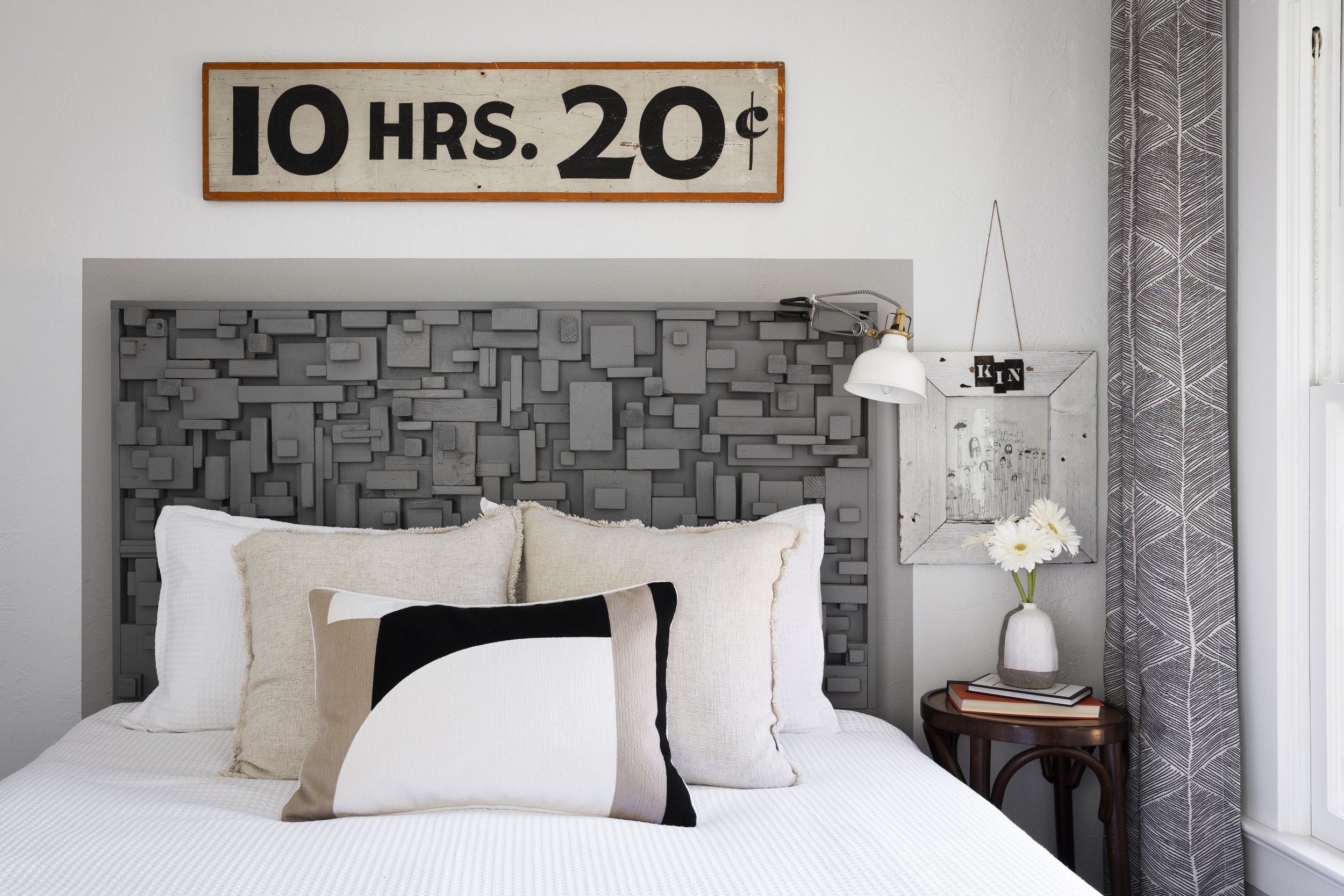 Bedroom with geometric headboard, vintage sign, pillows, side table with books, vase with daisies, and wall lamp.