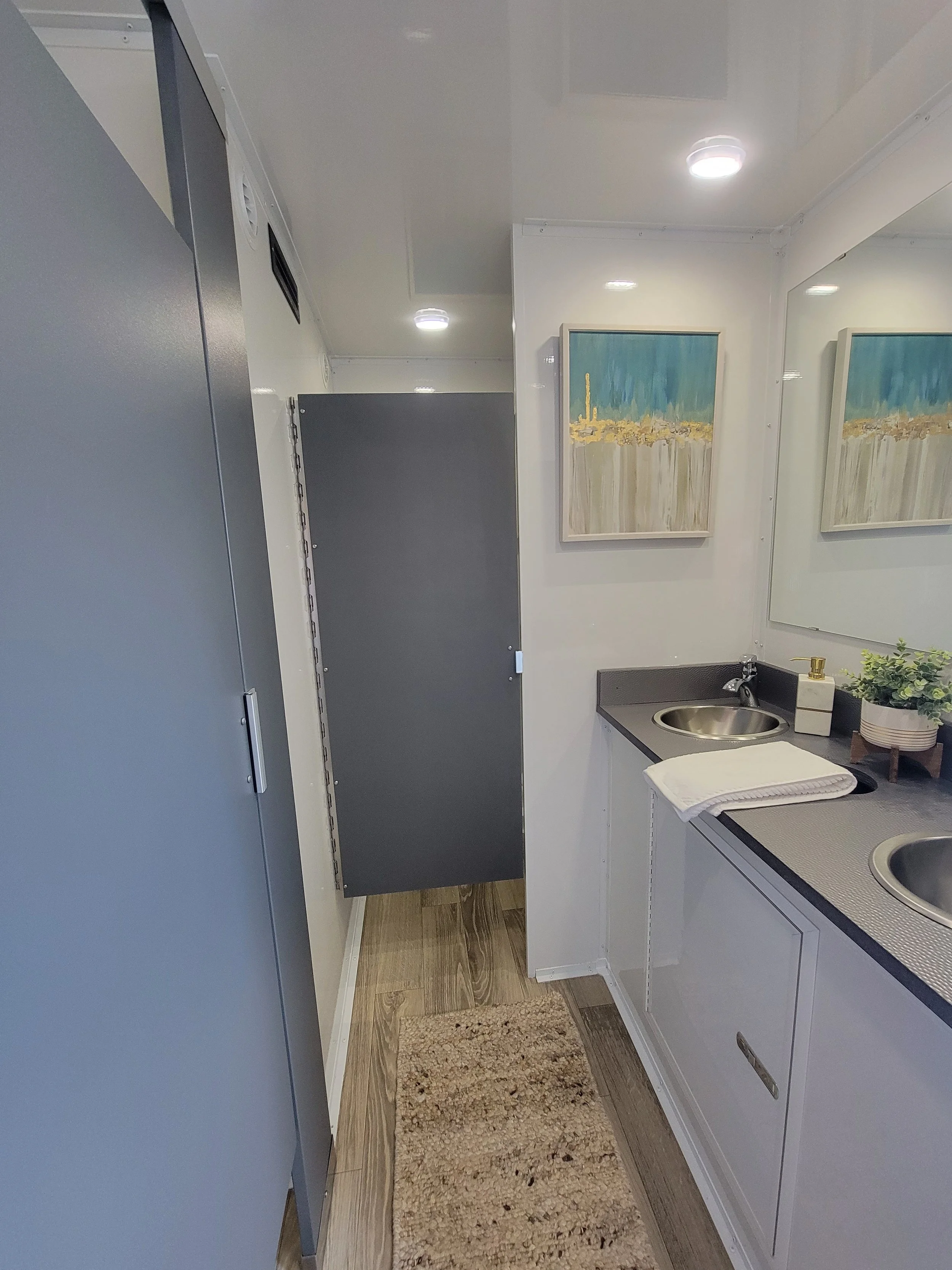 Interior of a modern restroom with gray stalls, dual sinks, and a decorative painting; features wood flooring and bright lighting.