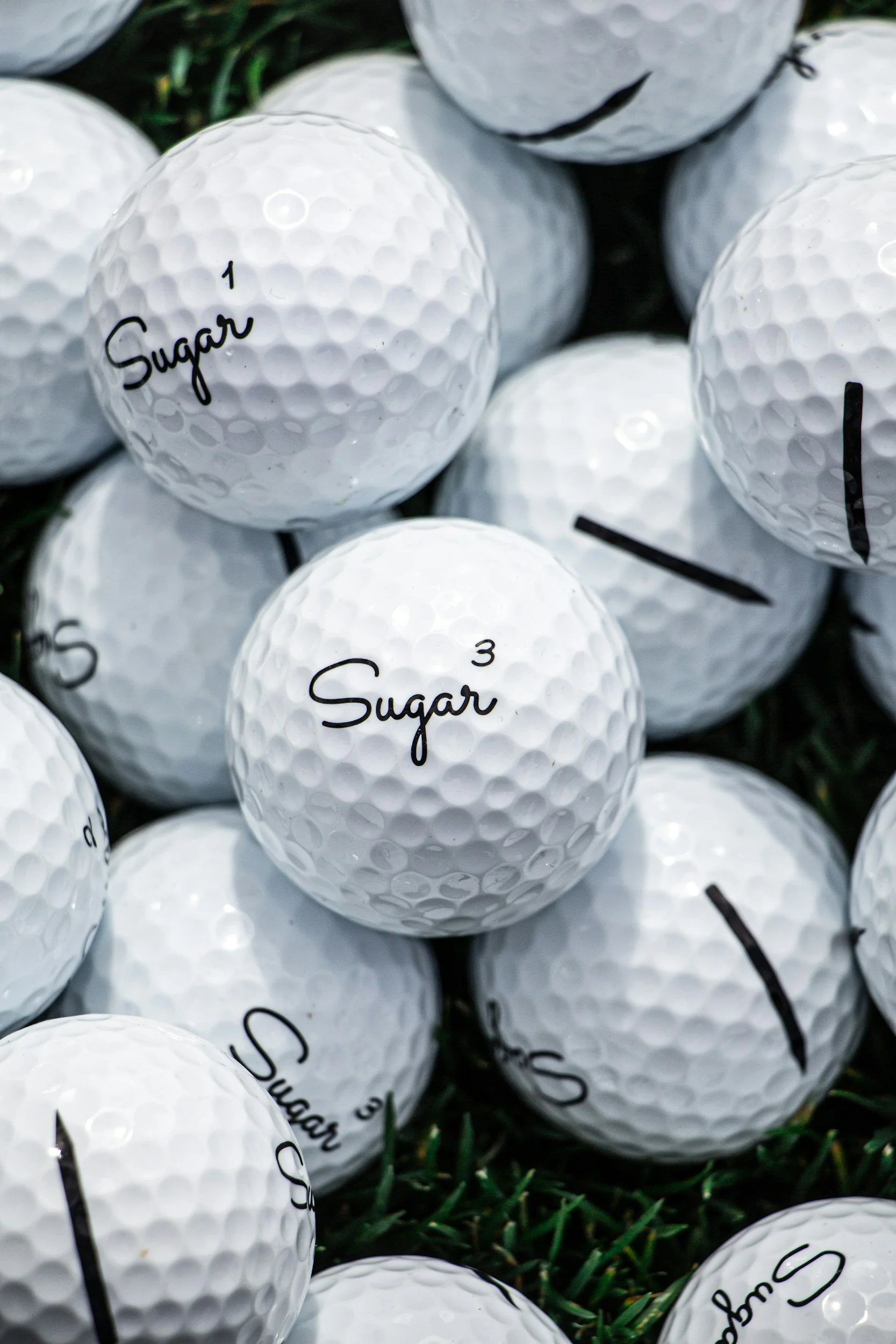 A close-up of multiple white golf balls on grass, with some marked 'Sugar' and numbered 1 and 3.