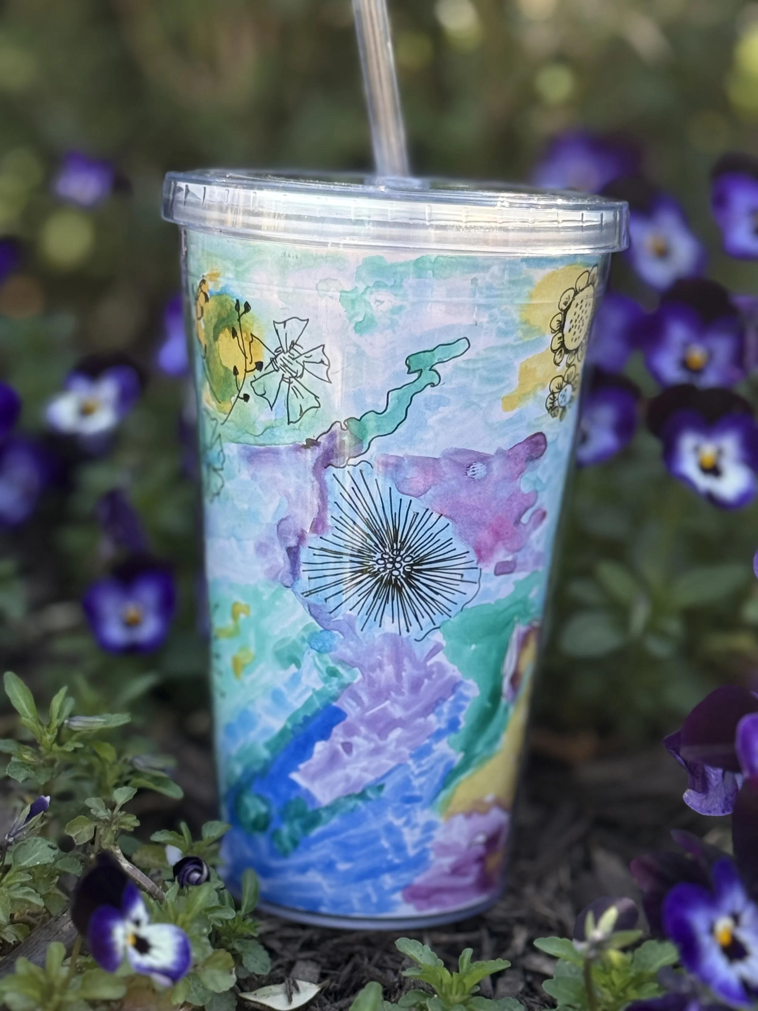 A colorful, artistic tumbler cup with a watercolor landscape design, including a mountain, sky, and flowers, placed among purple flowers outside.
