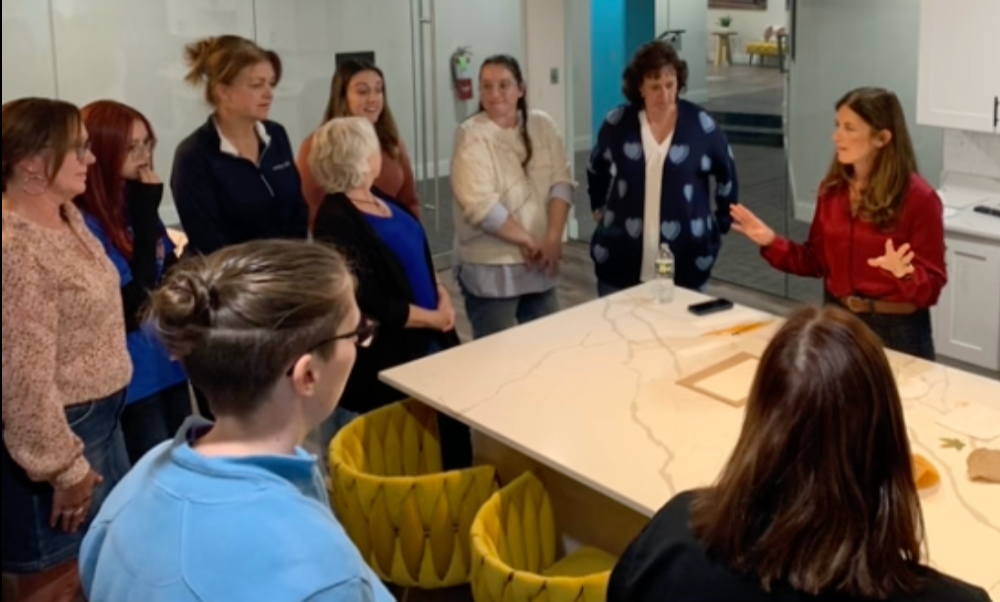 A woman in a red blouse is speaking to a group of women gathered around a large table with art supplies.