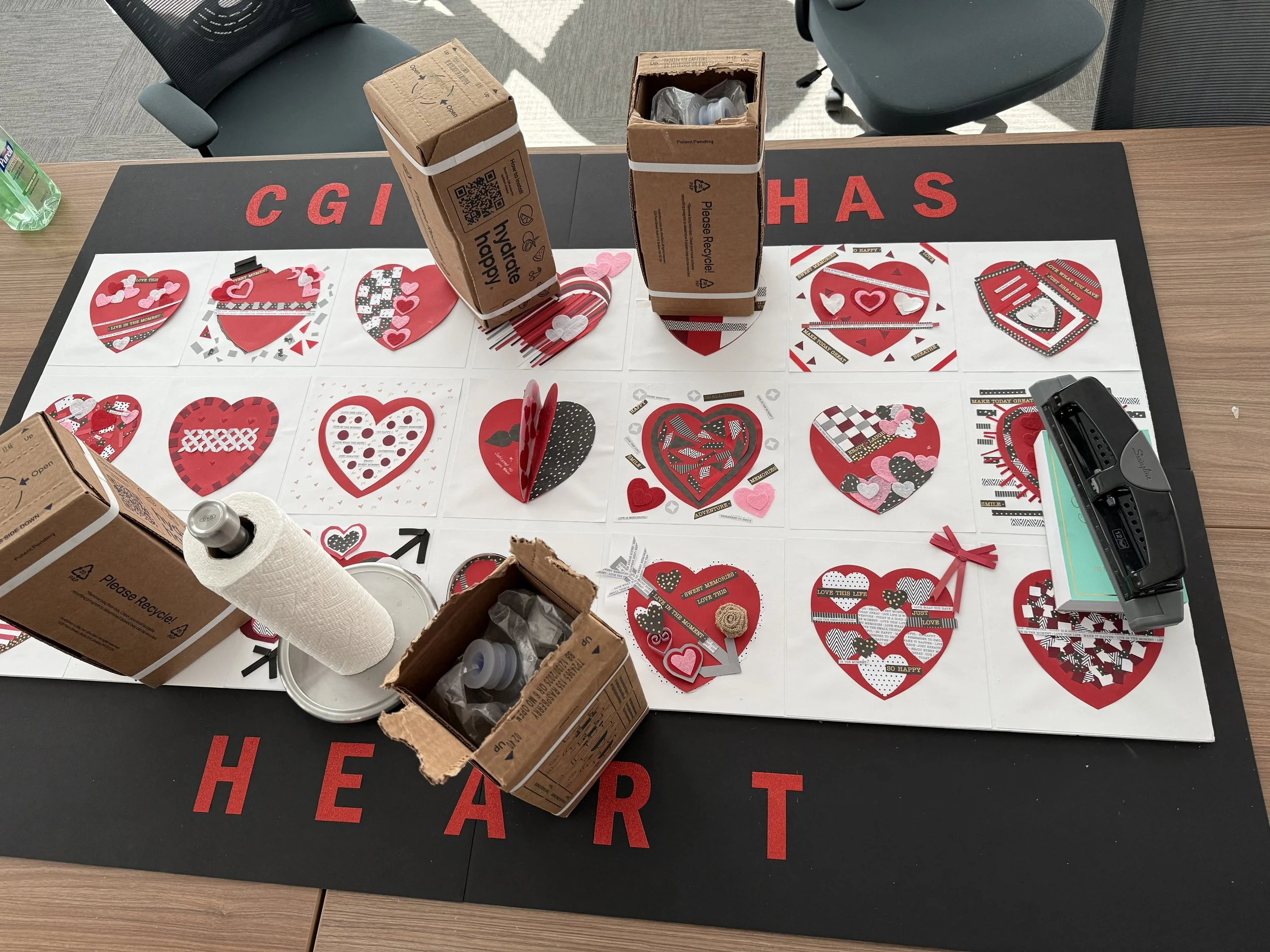A table with a Valentine-themed craft project, including red and black heart decorations, a paper towel roll, glue, and a device for cutting or punching paper. There are cartons and chairs in the background.