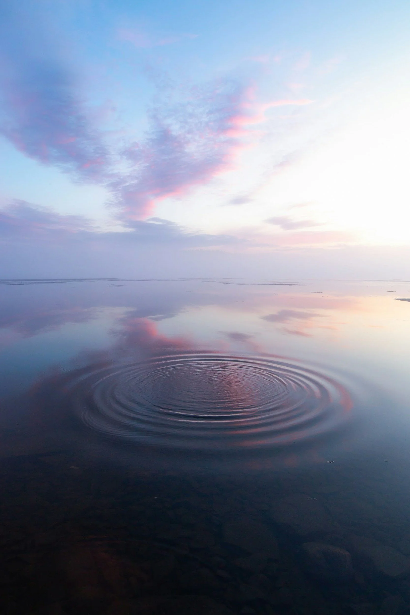 A tranquil body of water reflecting a sky with pink and purple clouds at sunset, with gentle ripples on the surface