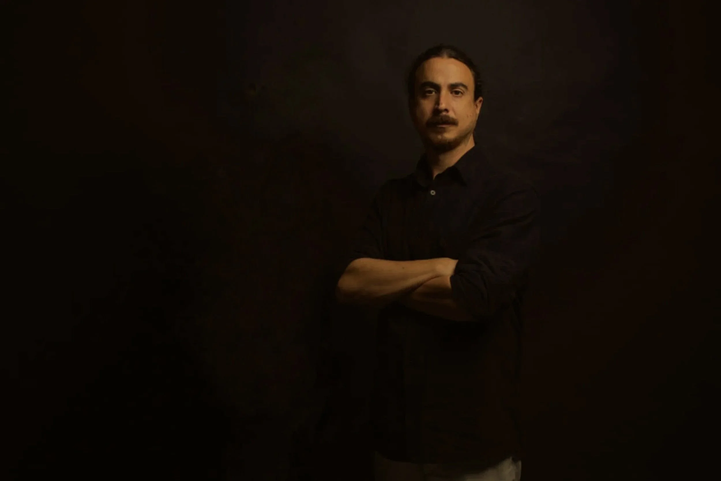 A man standing with arms crossed, wearing a black shirt, in a dimly lit environment with a dark background.
