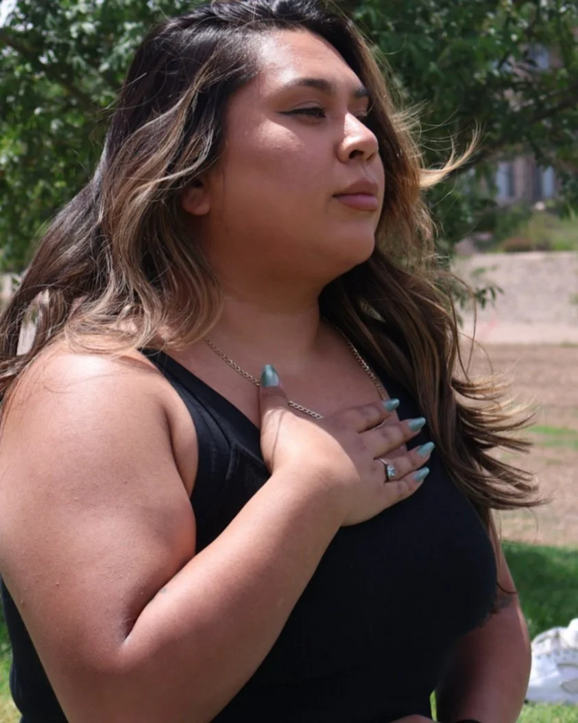 A woman with long brown hair and a black sleeveless top places her right hand over her chest, outdoors with green trees and a building in the background.