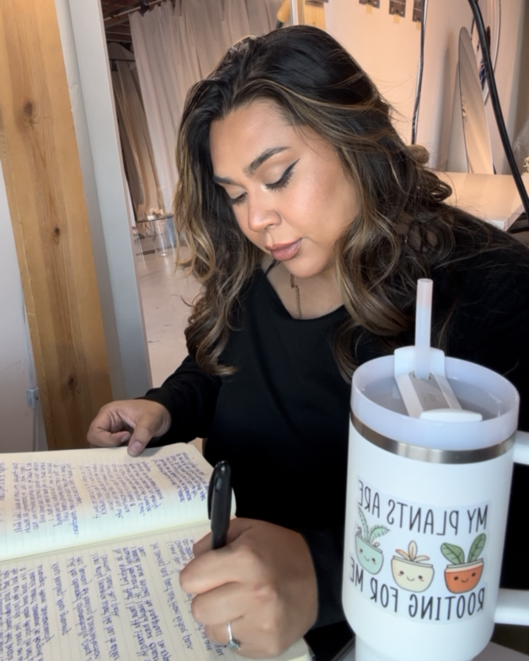 A woman with wavy, light brown hair and makeup, wearing a black long-sleeve top, is writing in a notebook with purple ink and a black pen. She is sitting at a table with a white tumbler that has a label reading 'My Plants Are Rooting For Me' and has a straw. The background shows a room with beige curtains and wooden accents.