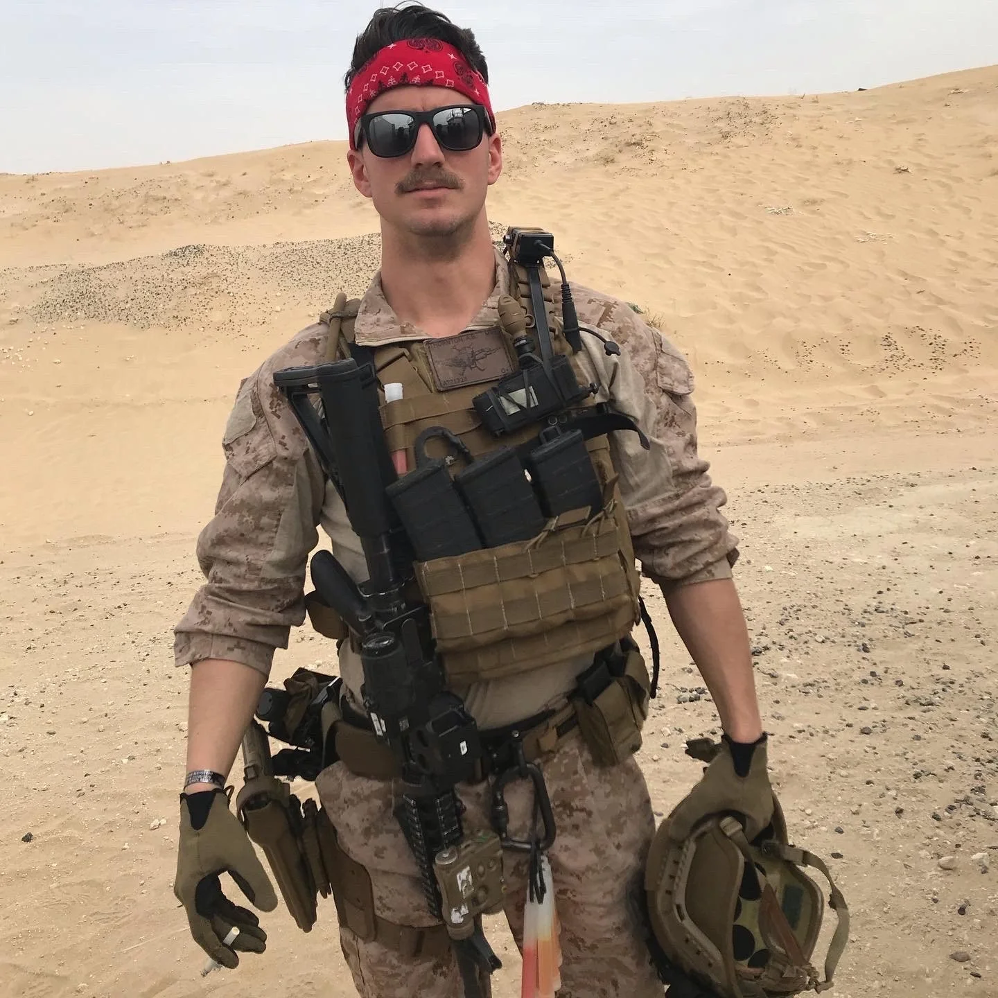 A man in desert camouflage military gear, wearing sunglasses and a red bandana, standing in a sandy desert landscape.