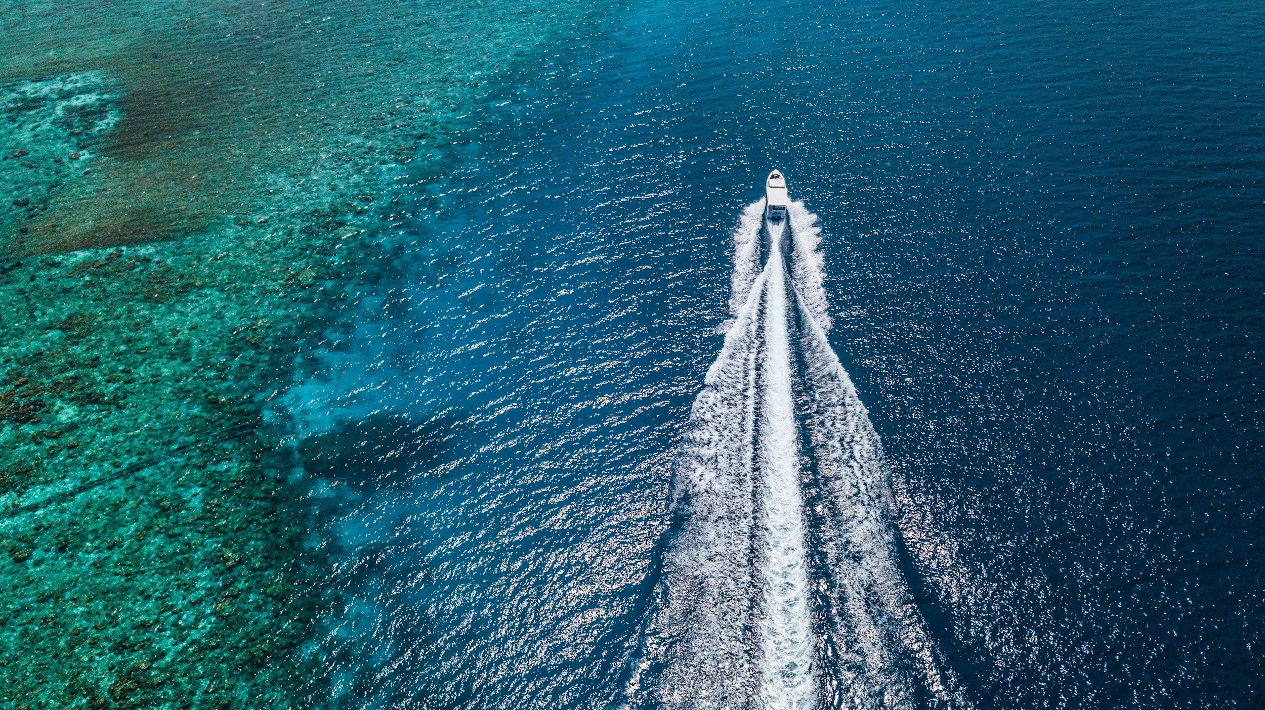 Aerial view of a white motorboat speeding through clear, deep blue water, creating a wake behind it. The water's color varies from turquoise near the shore to dark blue in the open sea.