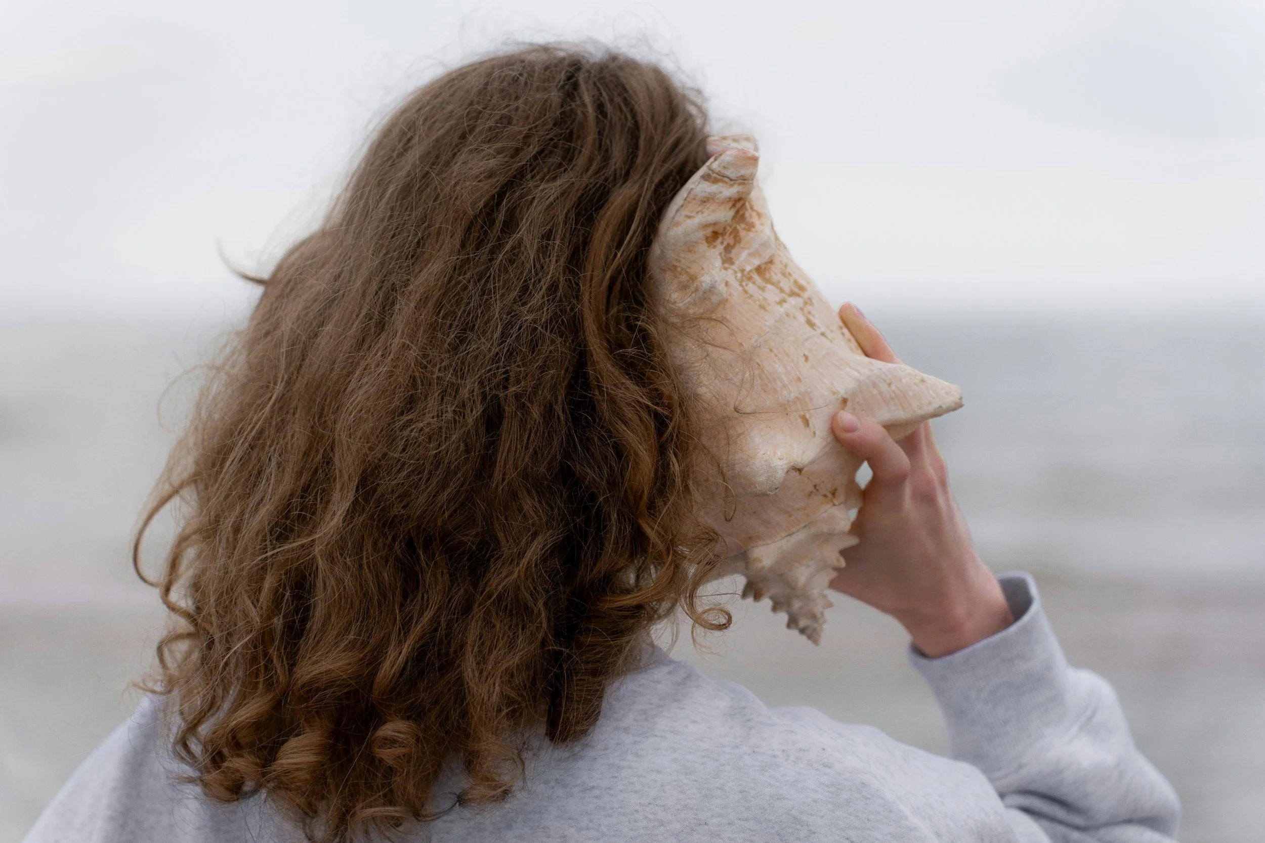 Person with curly hair holding a large seashell to their face, obscuring it, against a plain background.