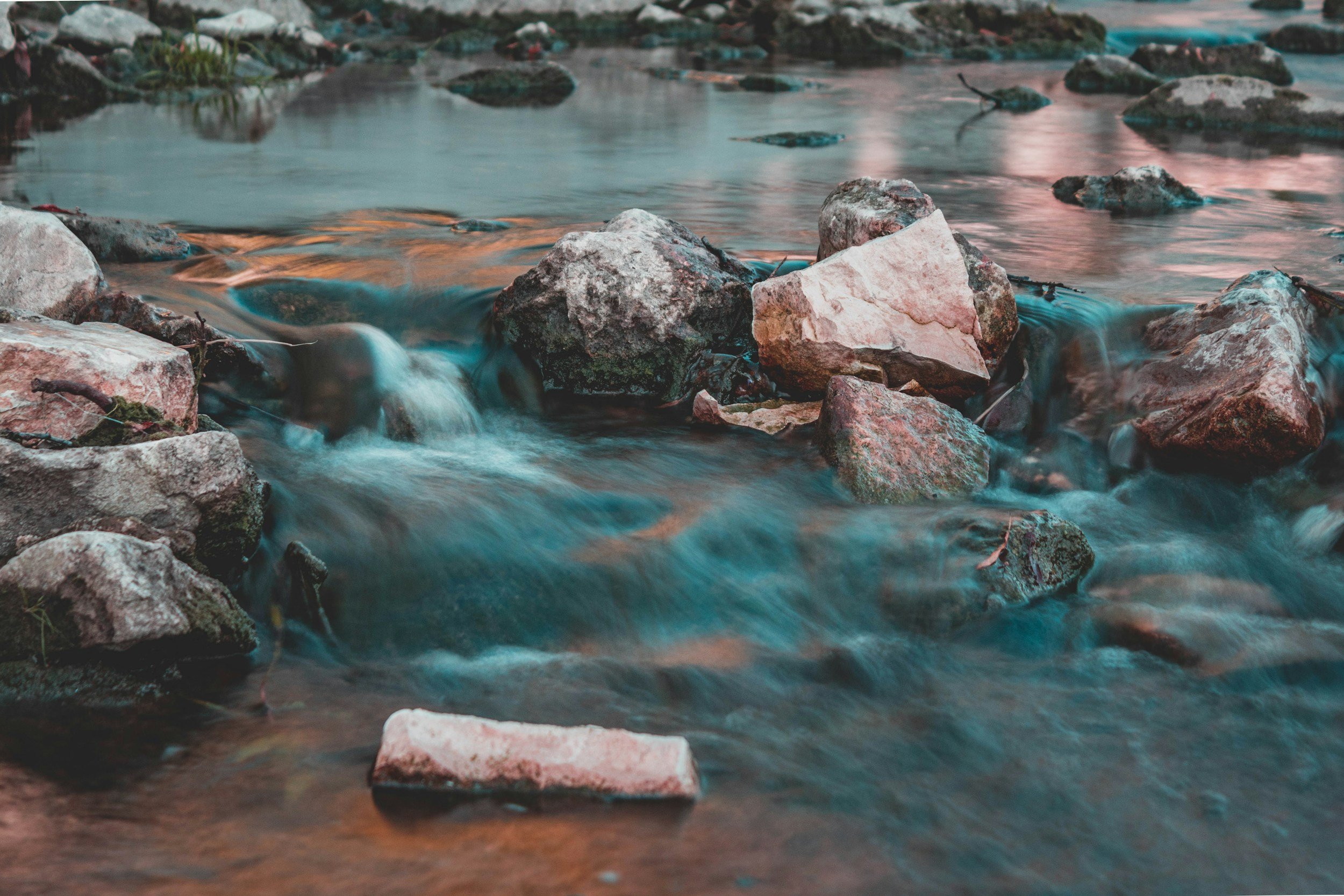 Close-up of a flowing river with rocks and small waterfalls, reflecting an orange and pink sunset sky.